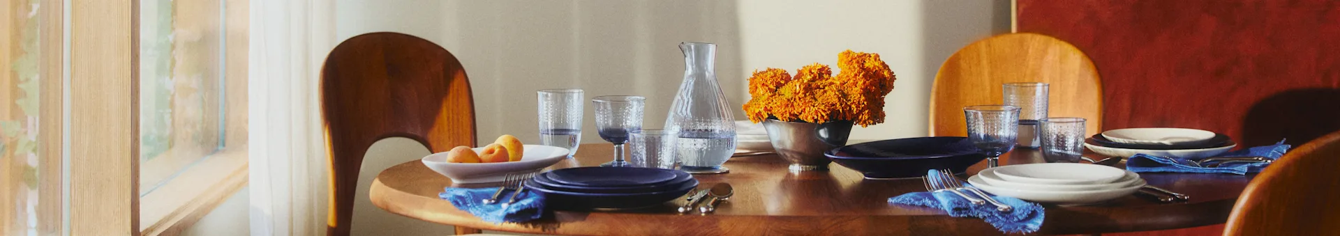 Table à manger ronde en bois avec assiettes bleues et blanches, serviettes bleues et fleurs oranges. La lumière du soleil filtre par une fenêtre.