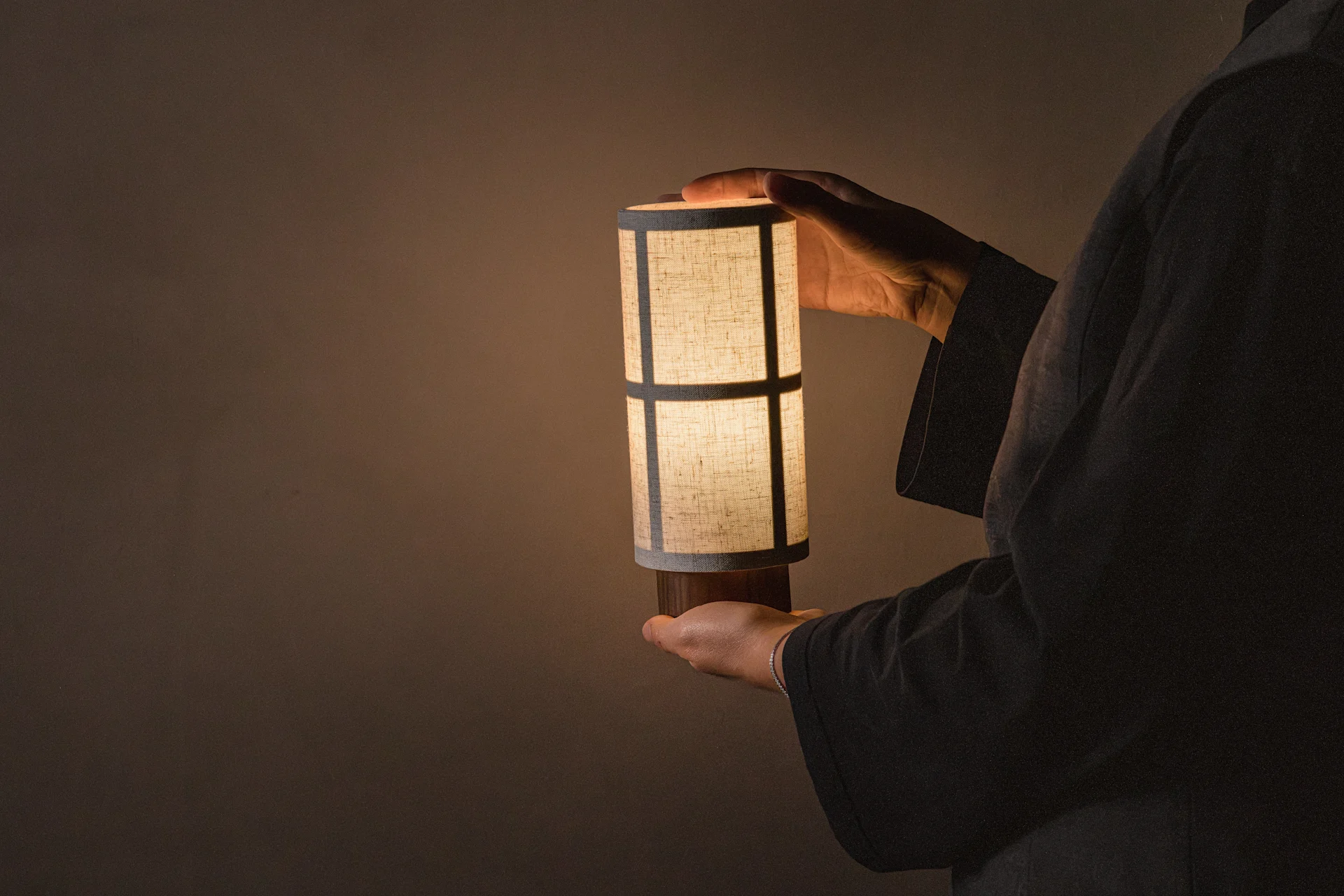 Personne tenant une lampe de table cylindrique éclairée avec un abat-jour en tissu à motifs, projetant une lueur chaude.