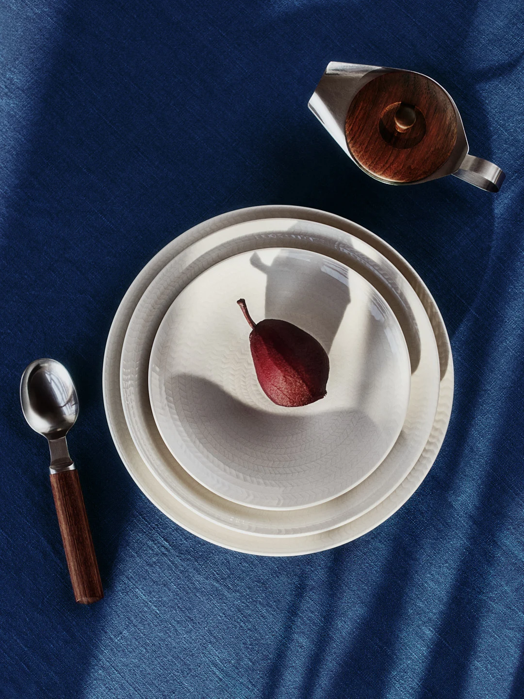 Poire rouge sur deux assiettes texturées, cuillère et crémier sur nappe bleue avec ombres.
