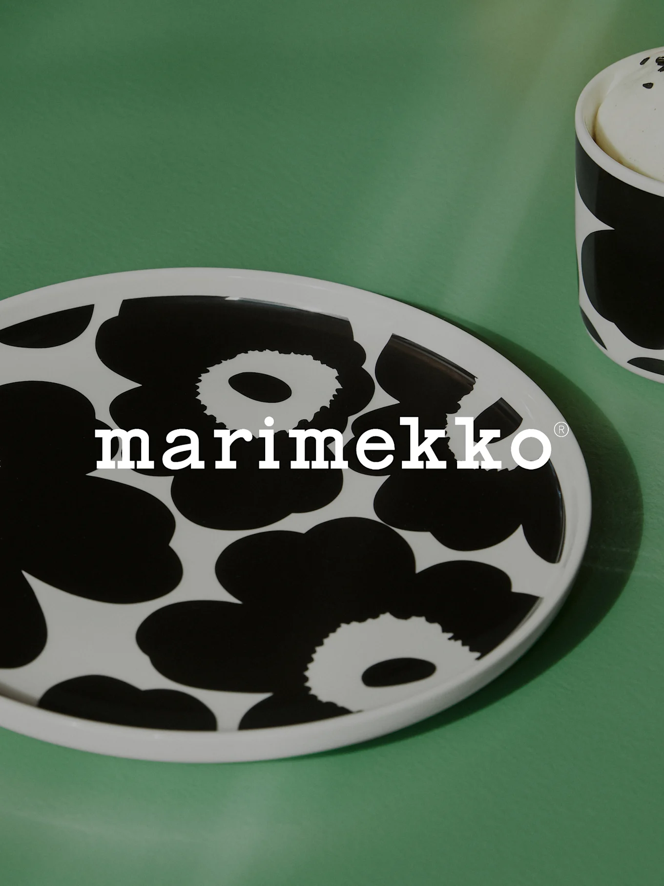 Assiette ovale blanche avec motif floral noir et logo Marimekko sur une surface verte, avec une tasse assortie partiellement visible.