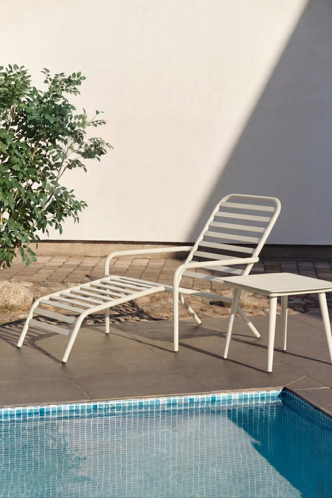 Chaise longue blanche et petite table d'appoint blanche au bord d'une piscine à l'eau turquoise. Plantes vertes et mur blanc en arrière-plan.