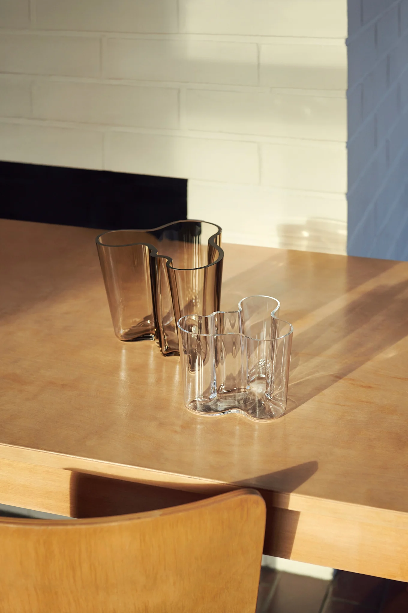 Trois vases en verre ondulés sur une table en bois. Deux sont ambrés, un est transparent, avec lumière du soleil et ombres sur la scène.