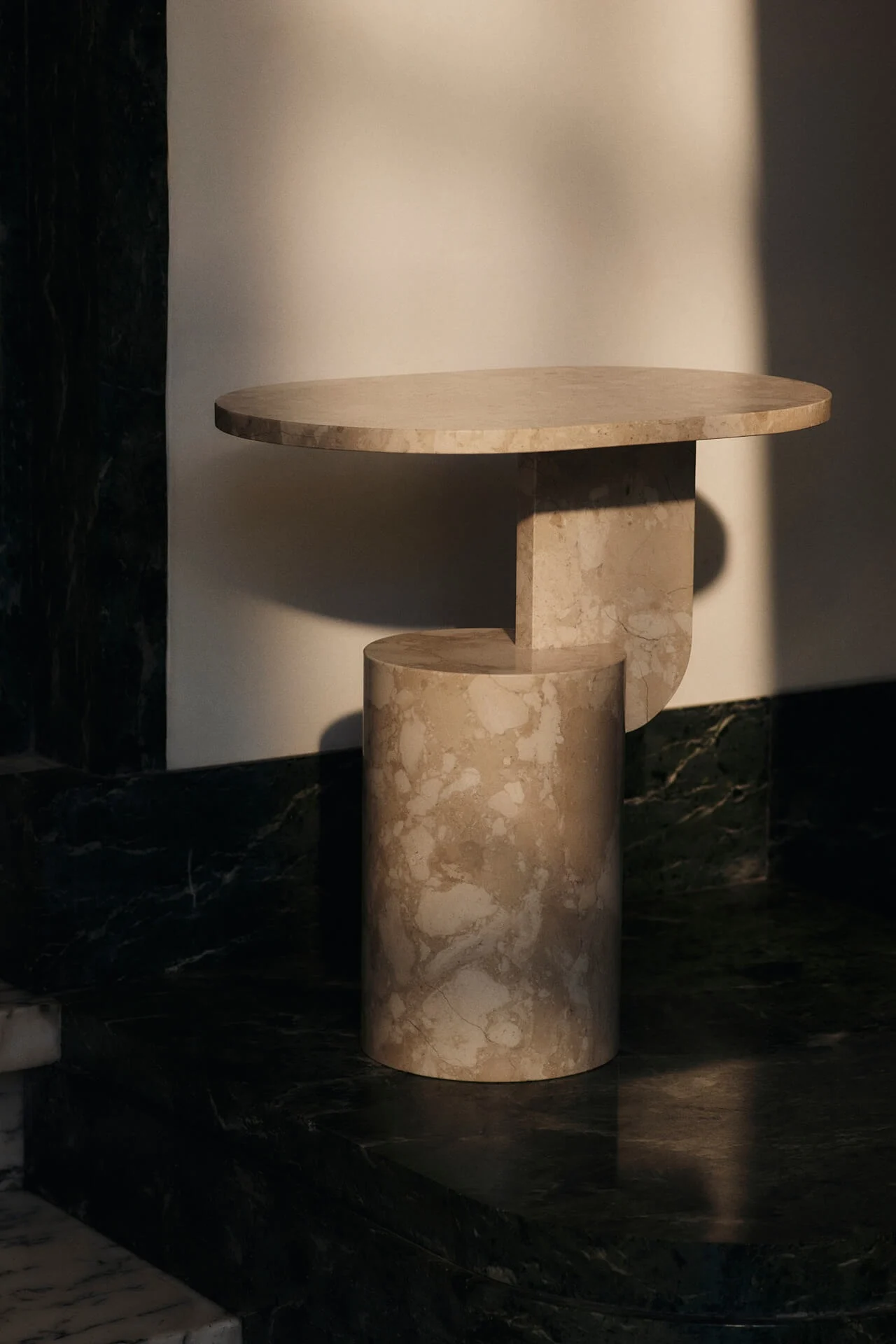 Table moderne en marbre beige, avec un plateau rond et un piètement géométrique, posée contre un mur clair.