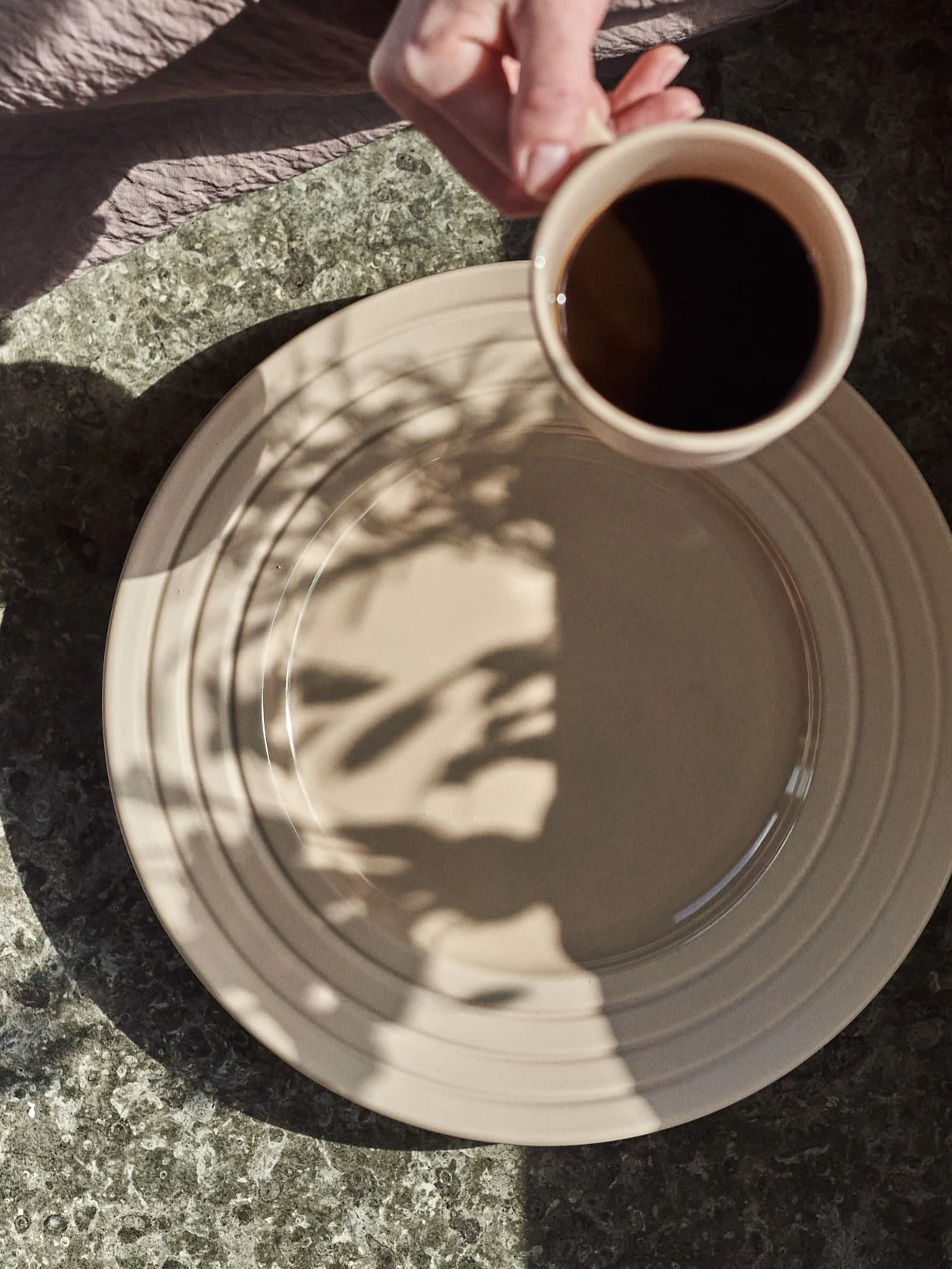 Main tenant une tasse de café au-dessus d'une assiette beige avec des ombres de feuilles sur une table mouchetée.