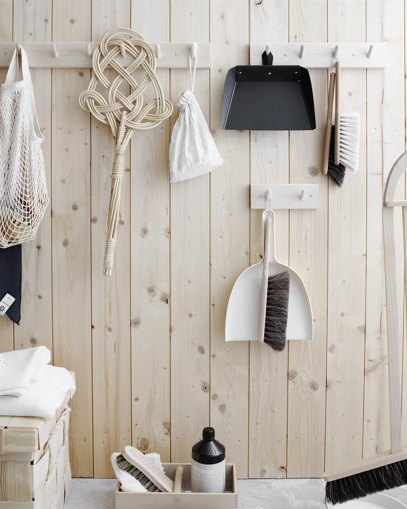 Outils de nettoyage et essentiels de lessive organisés sur un mur en bois clair. Comprend des brosses, des pelles, un batteur à tapis, un sac en filet, des serviettes et du détergent.
