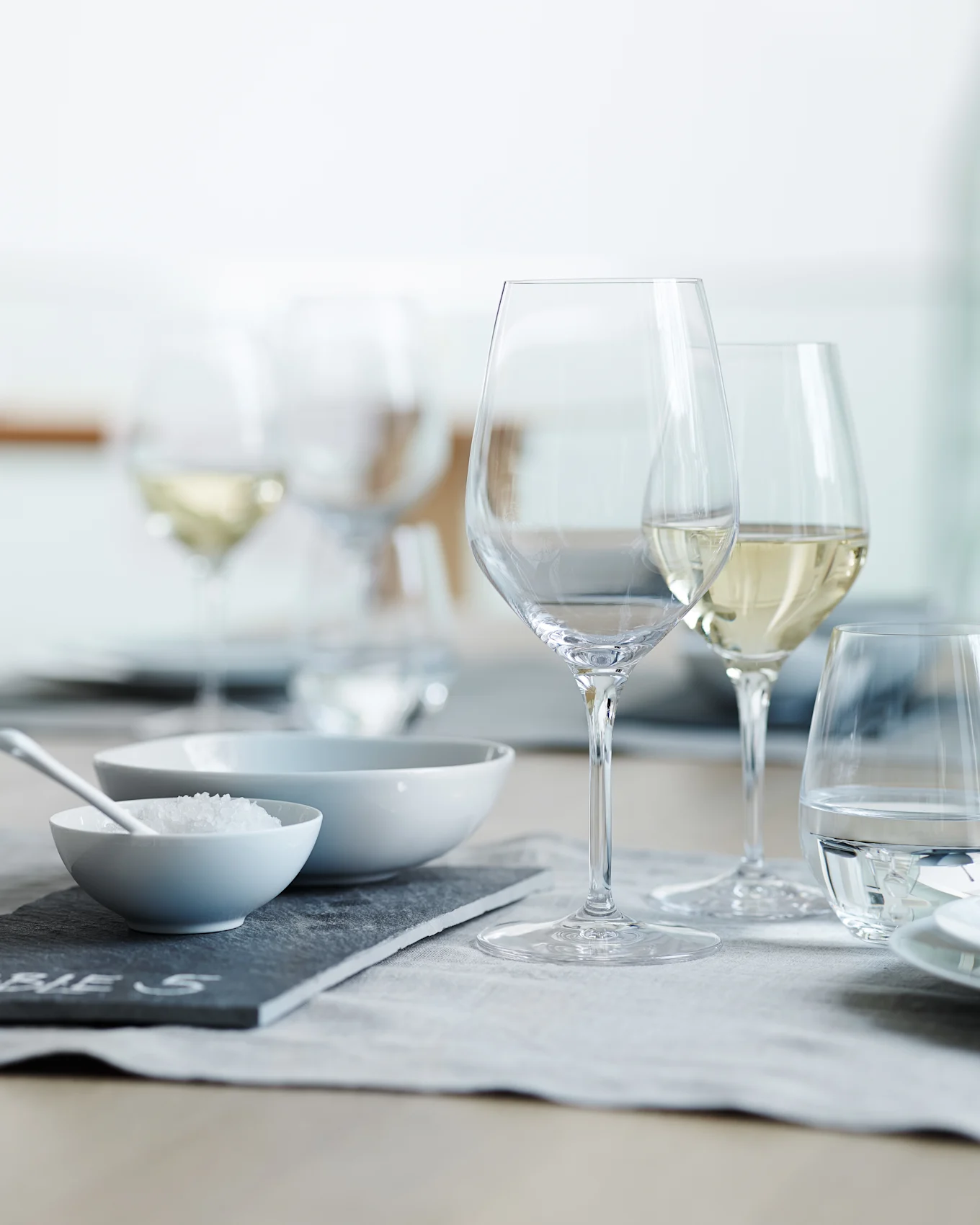 Une table dressée avec des verres à vin et à eau transparents, un bol de gros sel et un bol vide sur un set de table gris.