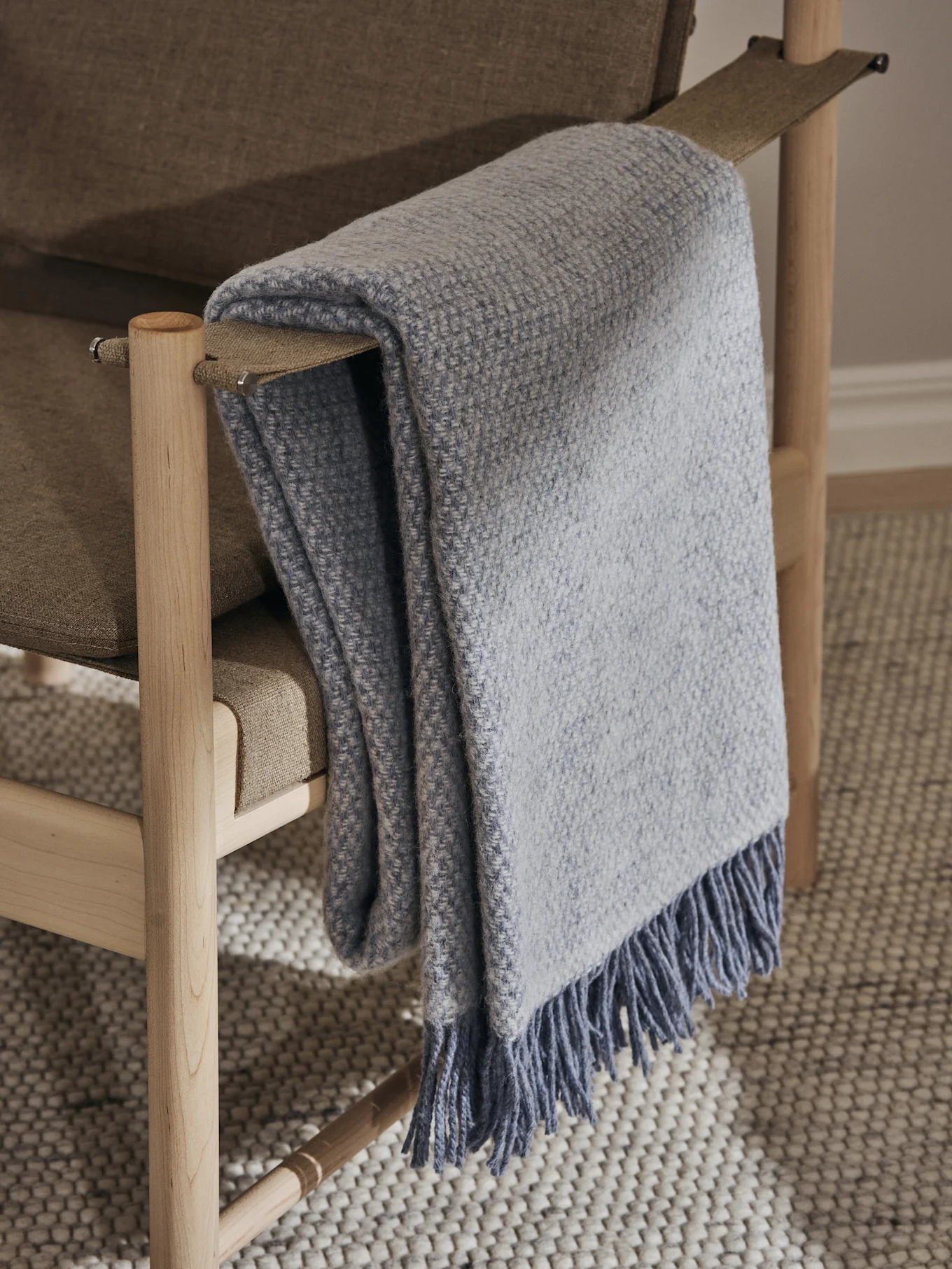 Plaid tissé bleu clair avec franges drapé sur un fauteuil rembourré marron clair avec cadre en bois clair, sur un tapis texturé.