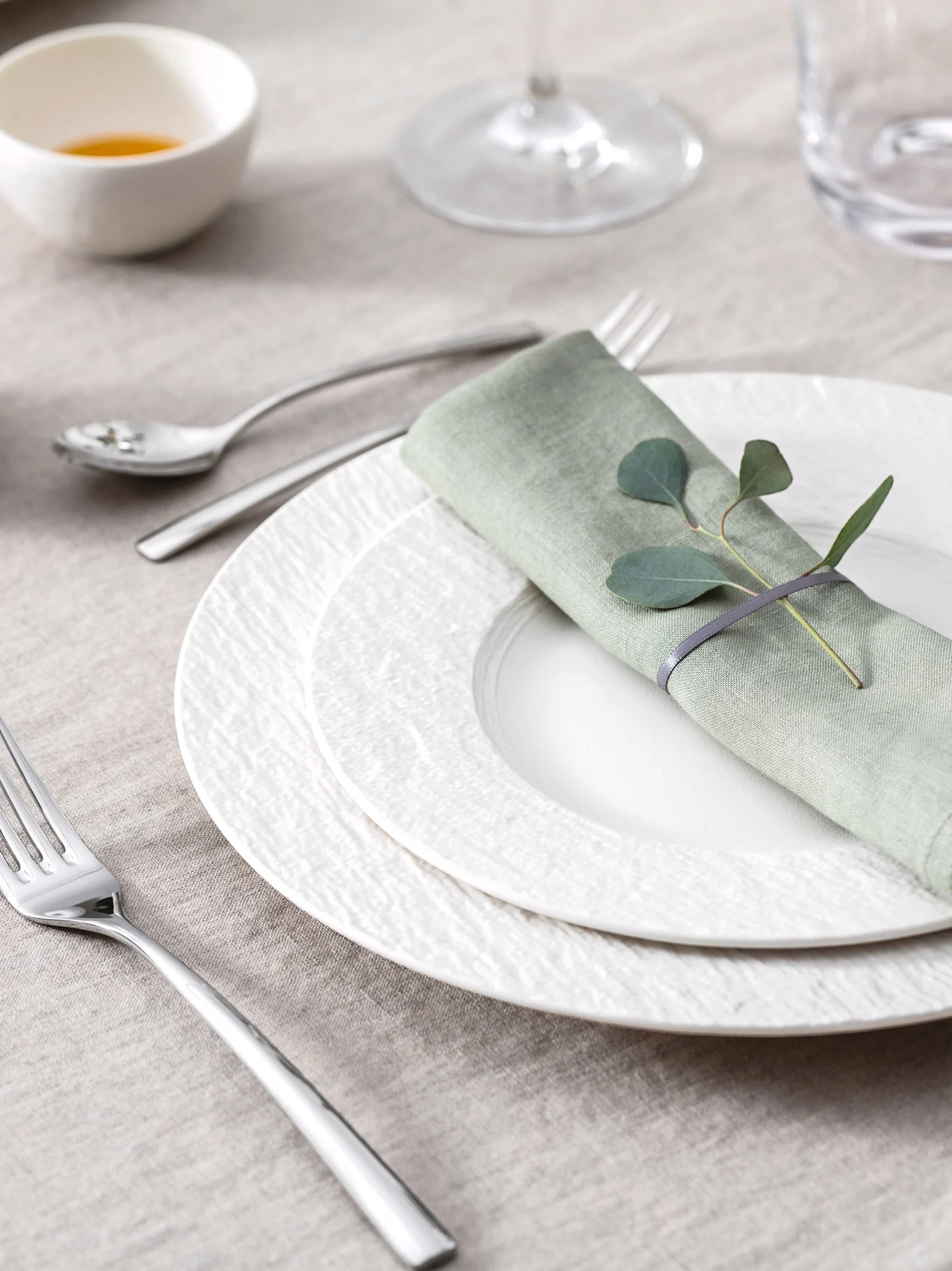 Élégante table dressée avec serviette en lin vert clair, feuilles d'eucalyptus, assiettes blanches texturées et couverts sur une nappe beige.