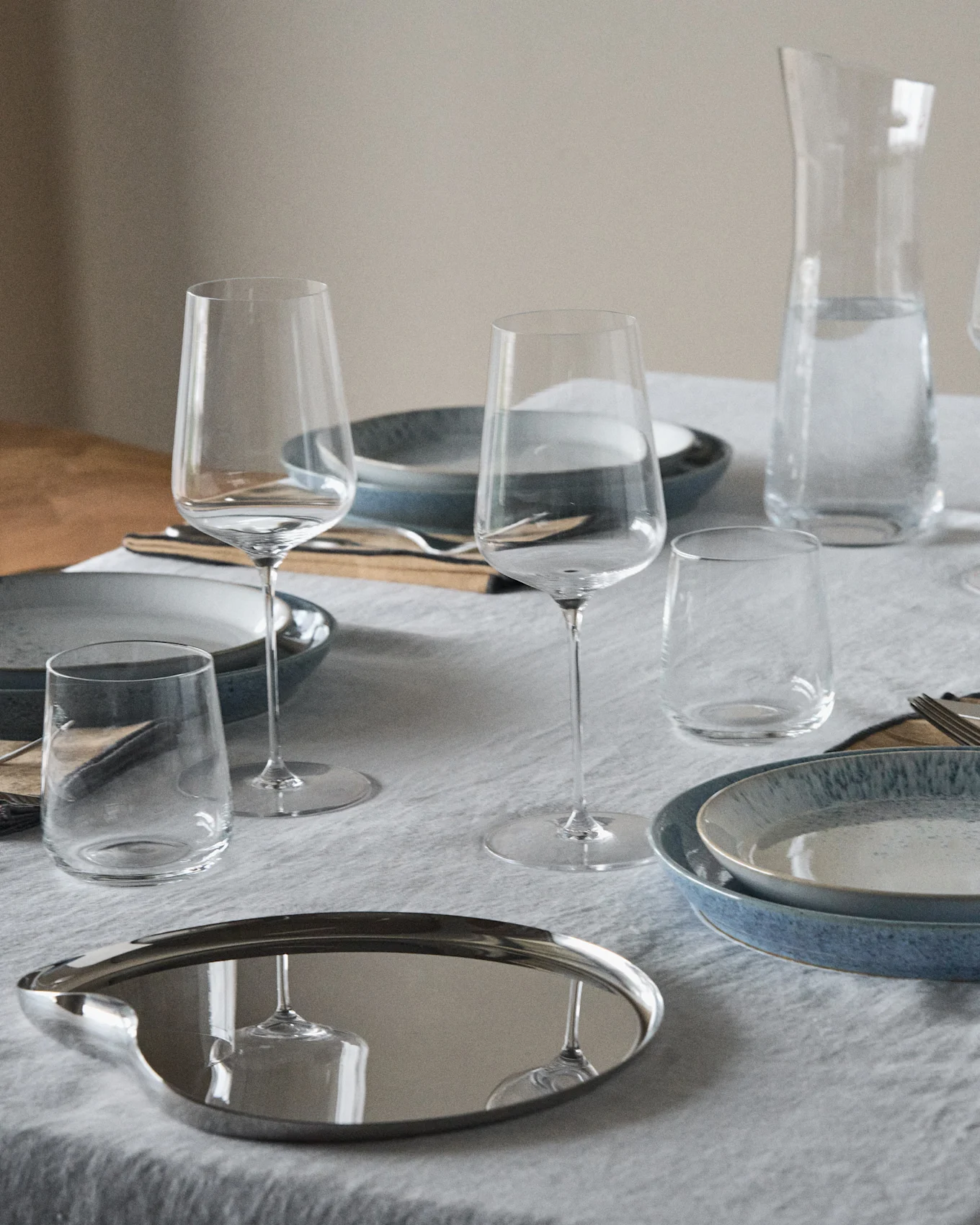 Table dressée avec des assiettes bleues et mouchetées, des verres à vin, des gobelets, une carafe d'eau et un plateau ovale réfléchissant.