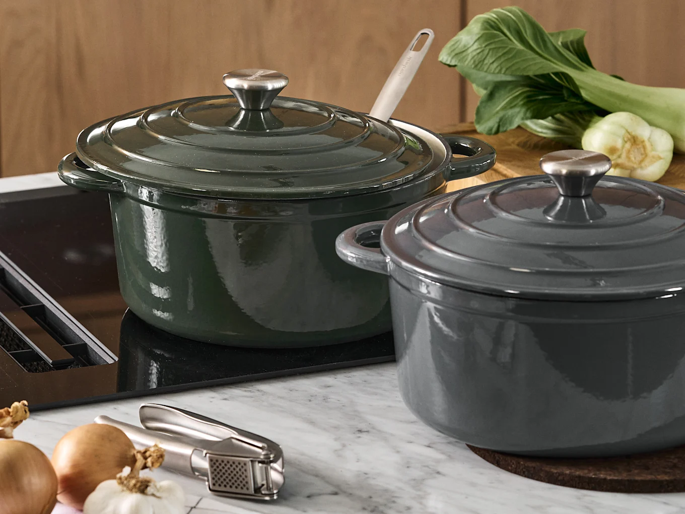 Deux cocottes en fonte avec couvercles, une vert foncé et une gris foncé, sur un comptoir de cuisine avec du bok choy, des oignons et un presse-ail.