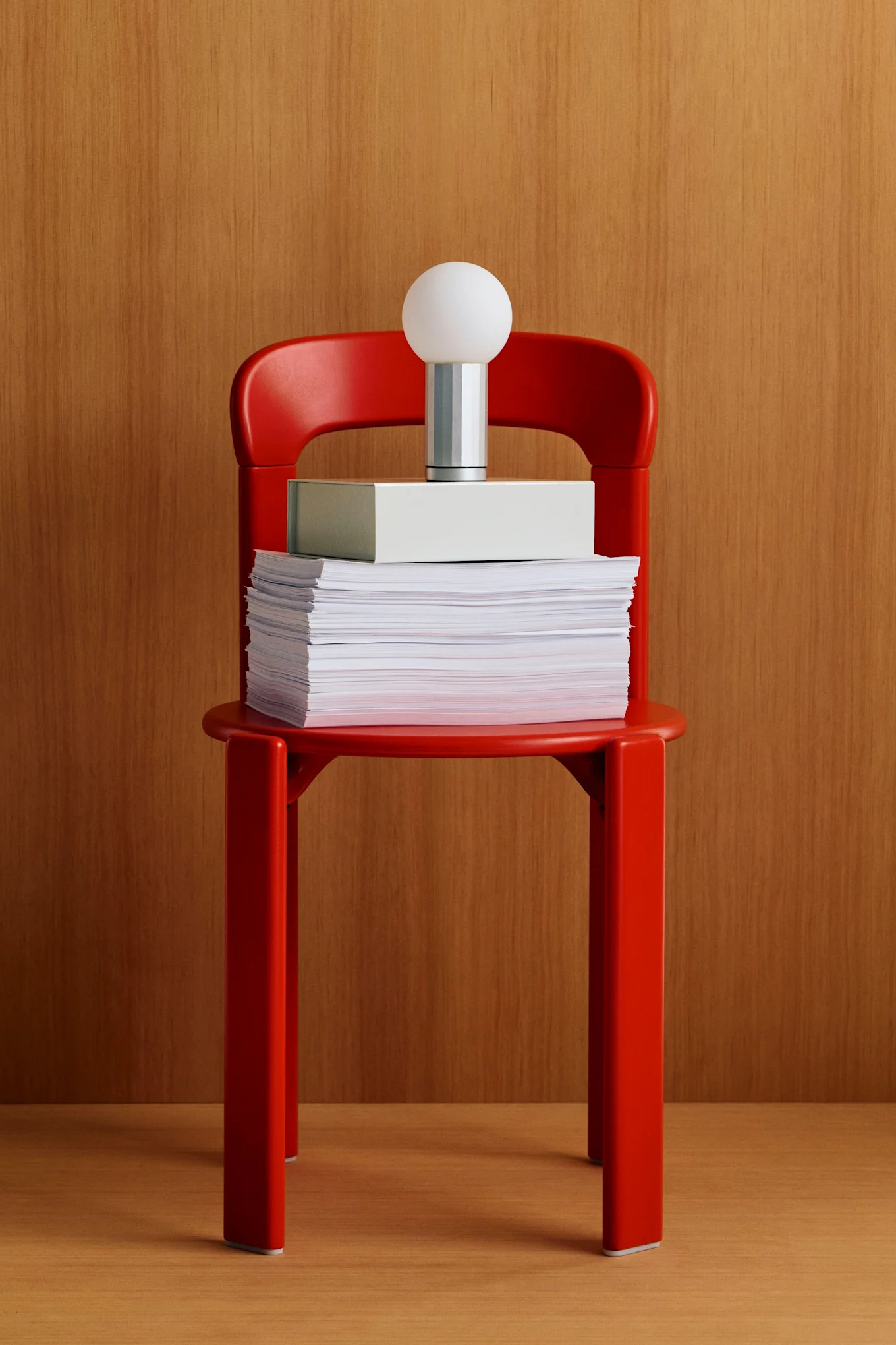 Chaise rouge contenant une pile de papiers, une boîte gris clair et une lampe moderne avec un abat-jour sphérique blanc sur fond en bois.