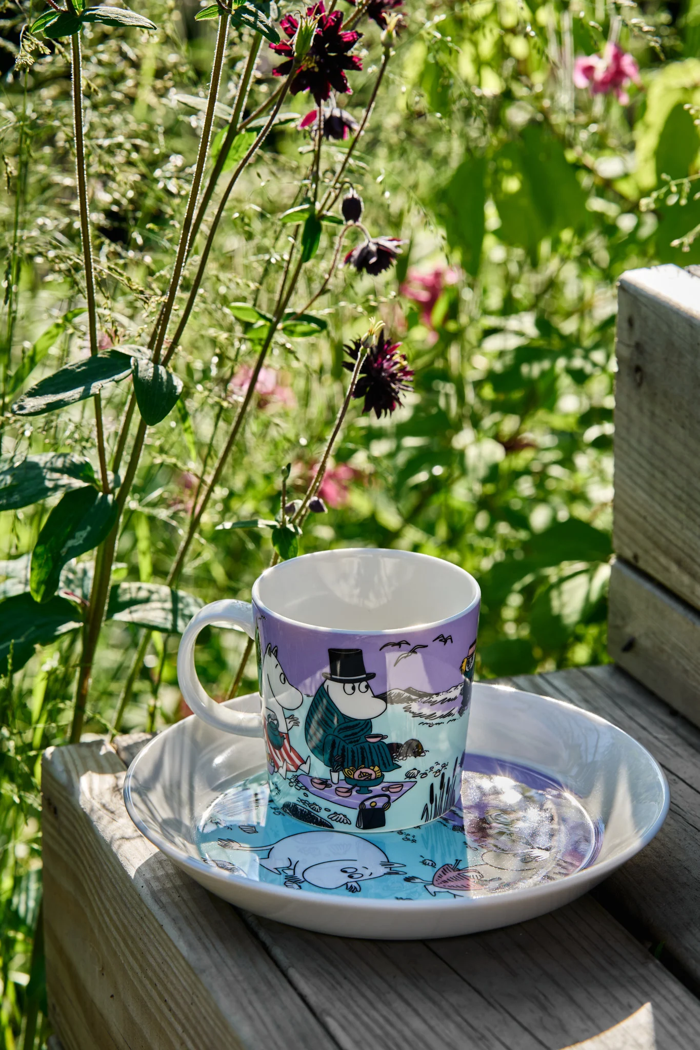 Le mug Moomin d'été 2025 « Beach Day » est posé sur une marche avec l'assiette assortie, avec une prairie sauvage en arrière-plan.