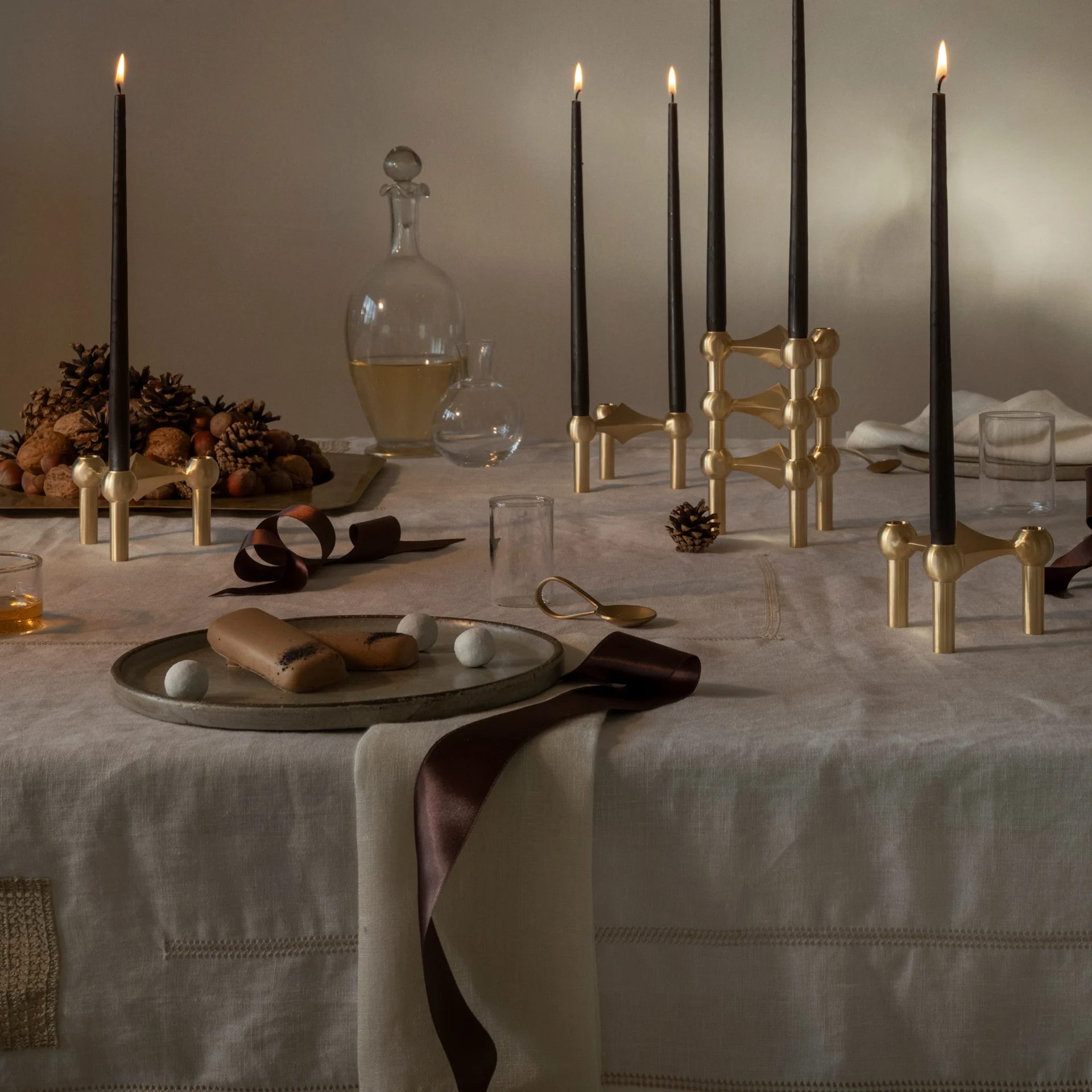 Table de fête avec bougies noires en bougeoirs modulaires laiton, panettone, noix, pommes de pin et desserts.