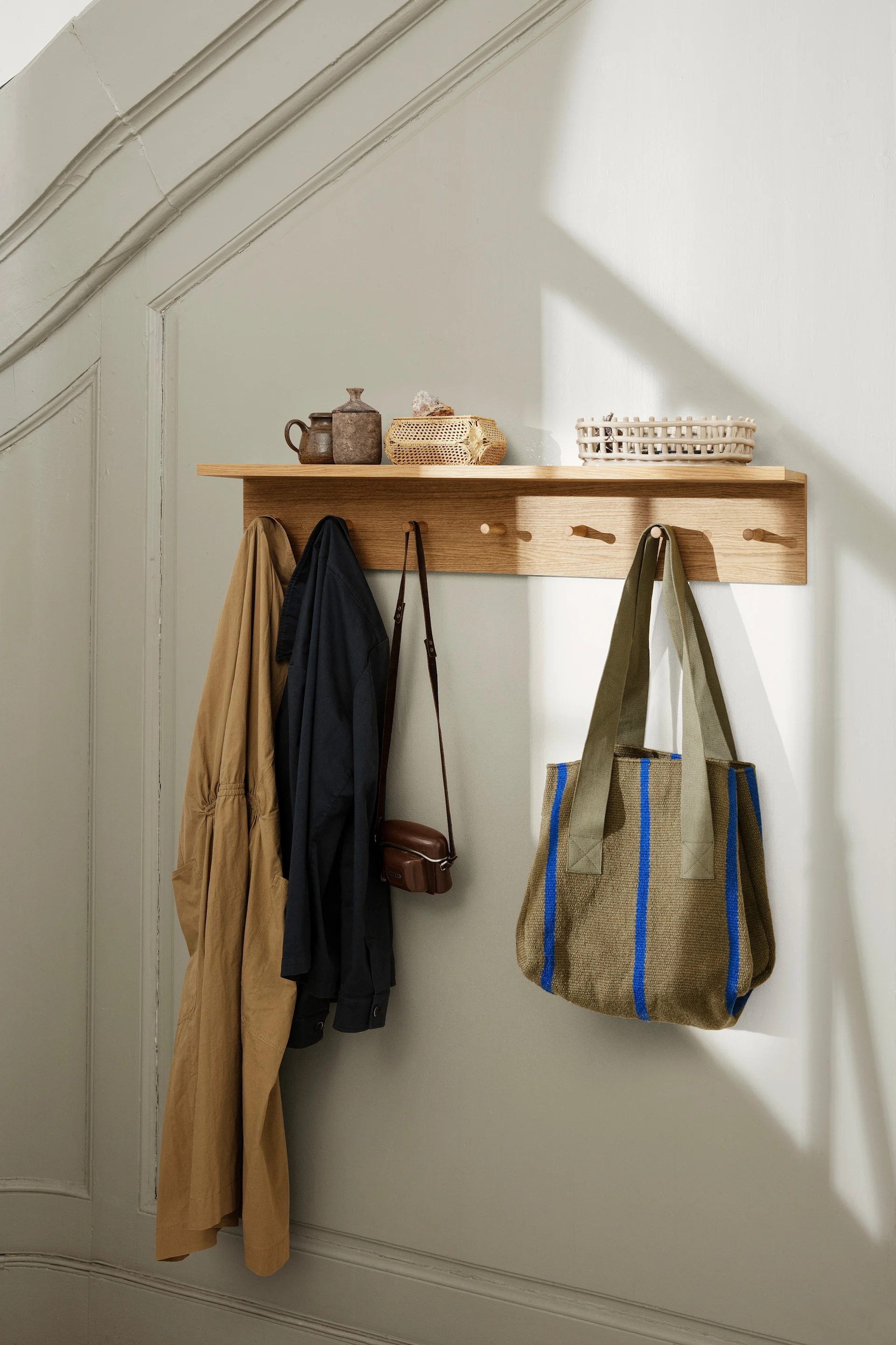 Étagère murale en bois avec patères. Deux vestes, petit sac en cuir, cabas vert olive à rayures bleues. Pots et paniers dessus.