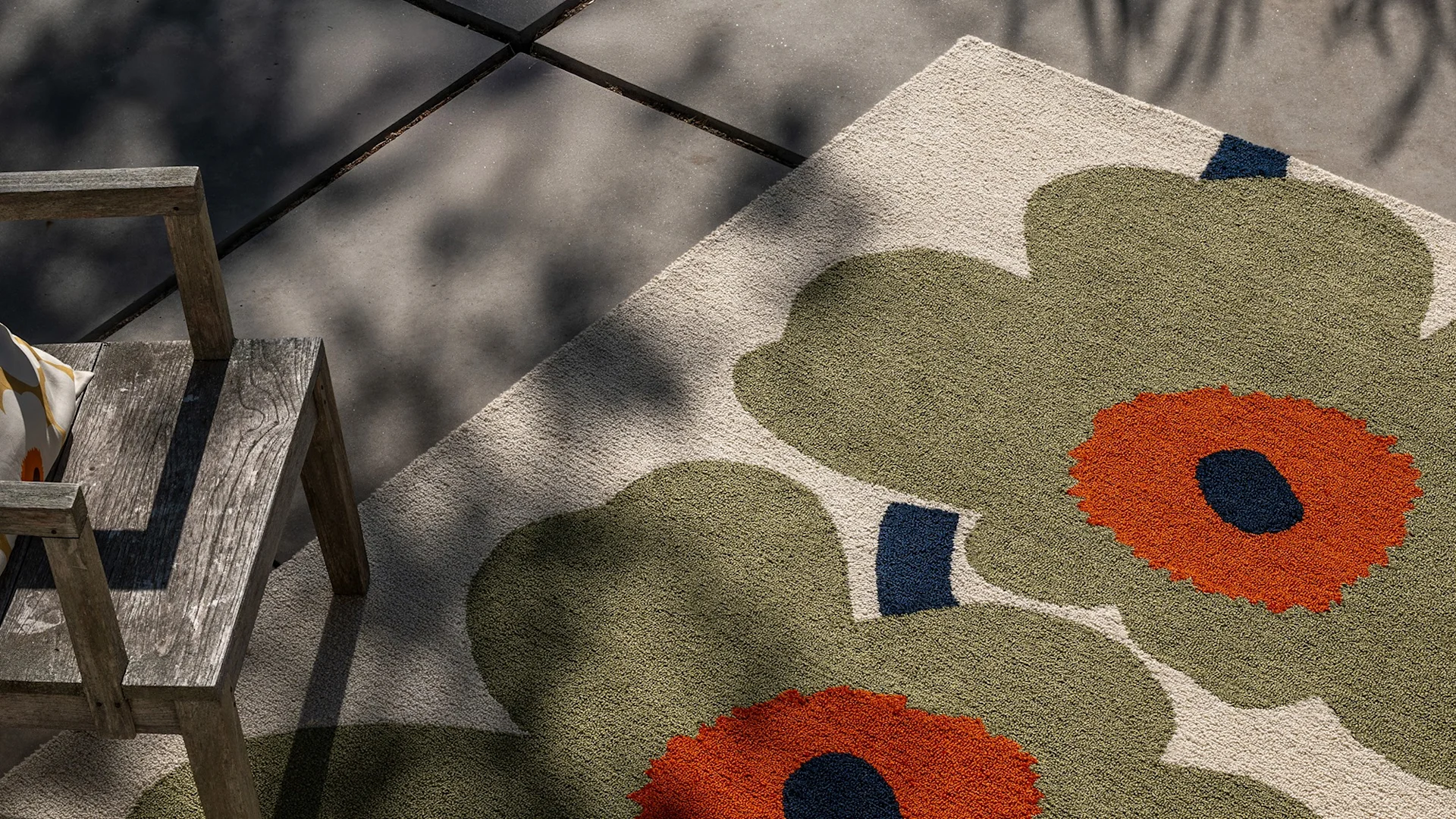 Vue plongeante d'un tapis d'extérieur avec de grands motifs floraux vert olive et orange sur fond crème, à côté d'une chaise en bois sur un patio en béton.