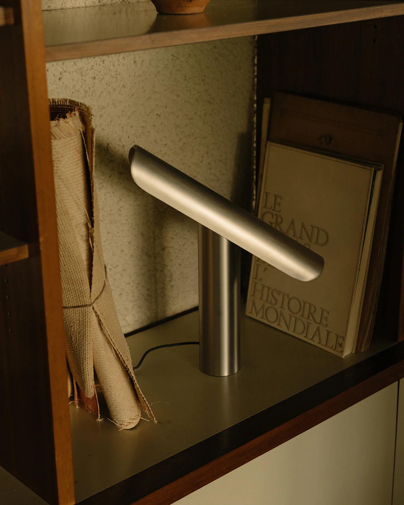 Lampe de bureau argentée moderne avec une base cylindrique et une barre lumineuse horizontale inclinée sur une étagère.
