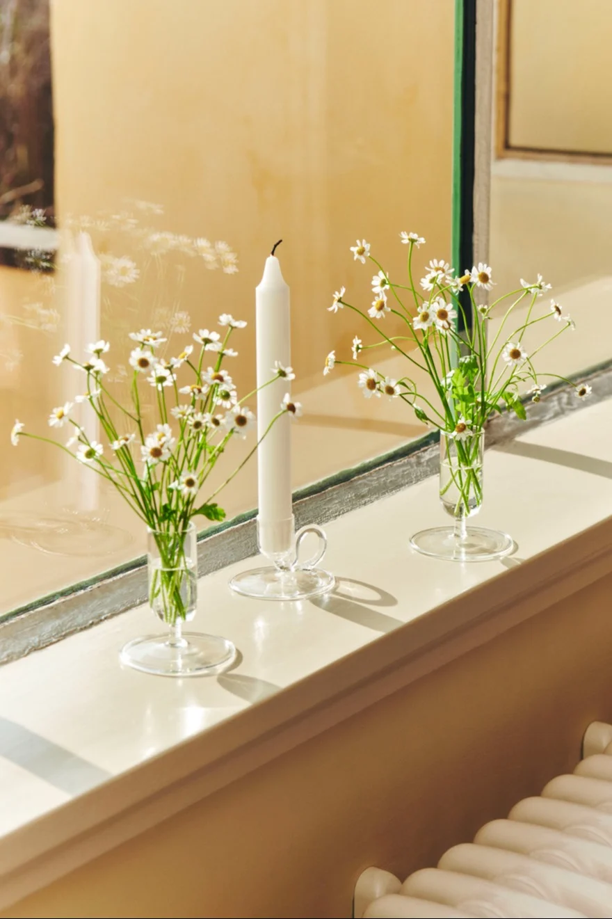 Trois objets décoratifs en verre transparent sur un rebord de fenêtre ensoleillé : deux vases avec des marguerites blanches et une grande bougie blanche dans un bougeoir.
