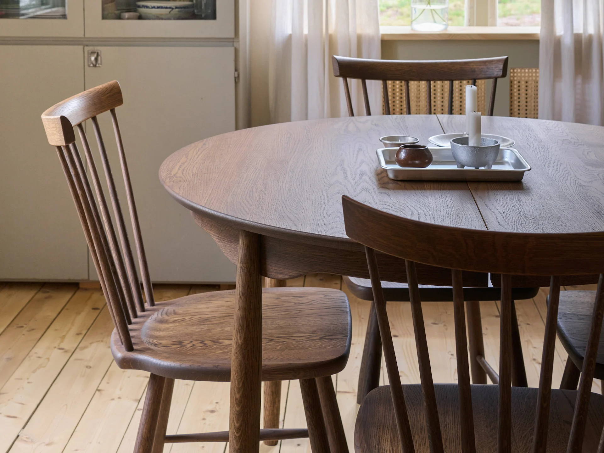 Une table à manger ronde en bois avec trois chaises à dossier à barreaux, un plateau avec des bougies et un bol, et un plancher en bois.