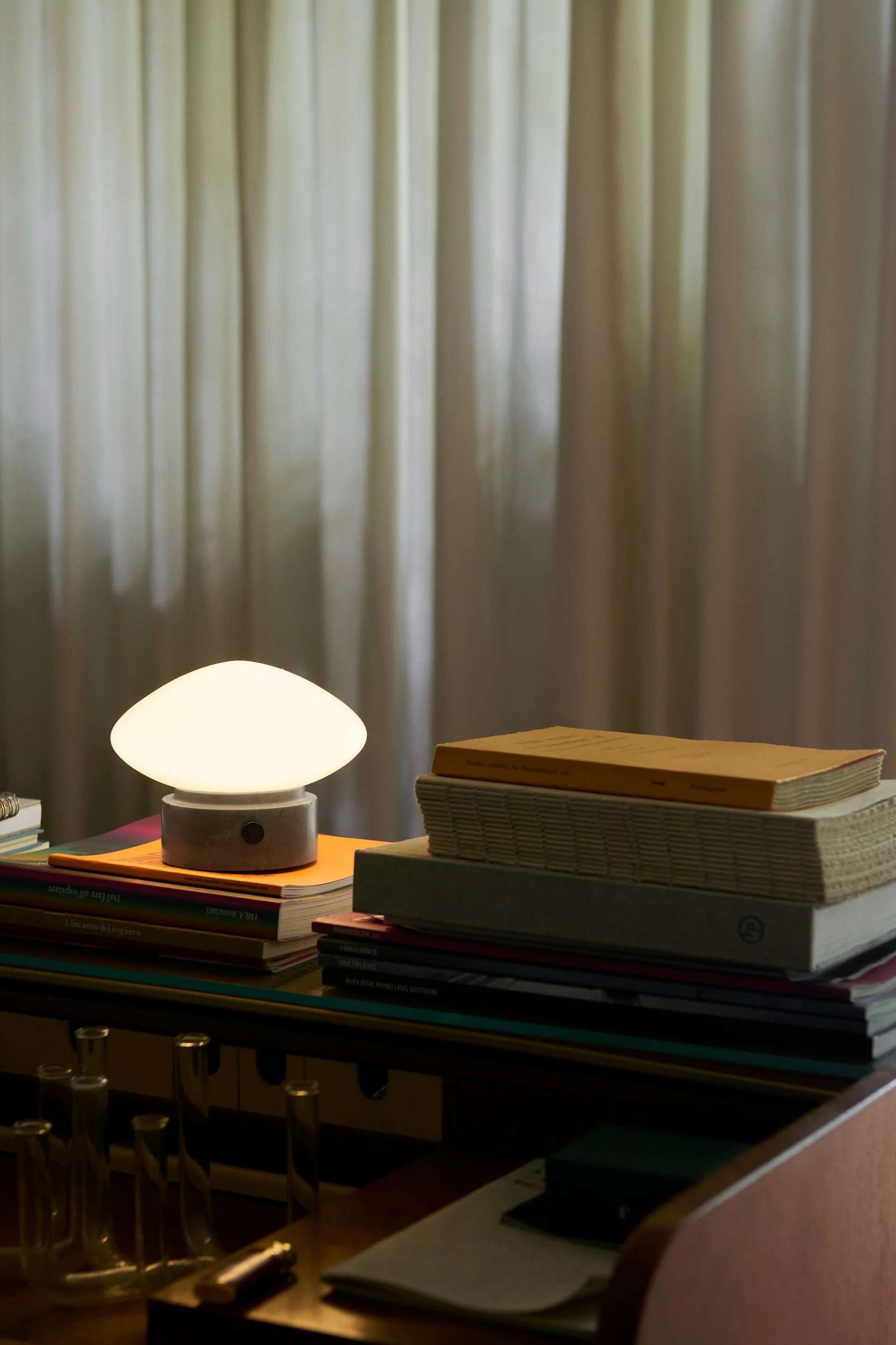 Lampe de table champignon avec dôme blanc lumineux et base grise, posée sur des livres sur un bureau.