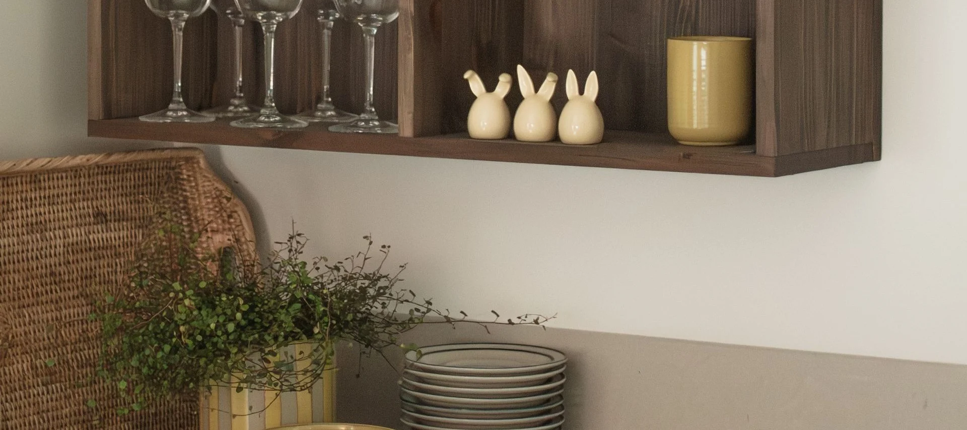 Une étagère en bois avec verres, trois figurines de lapins et une tasse jaune, avec un panier en osier et des plantes en dessous.