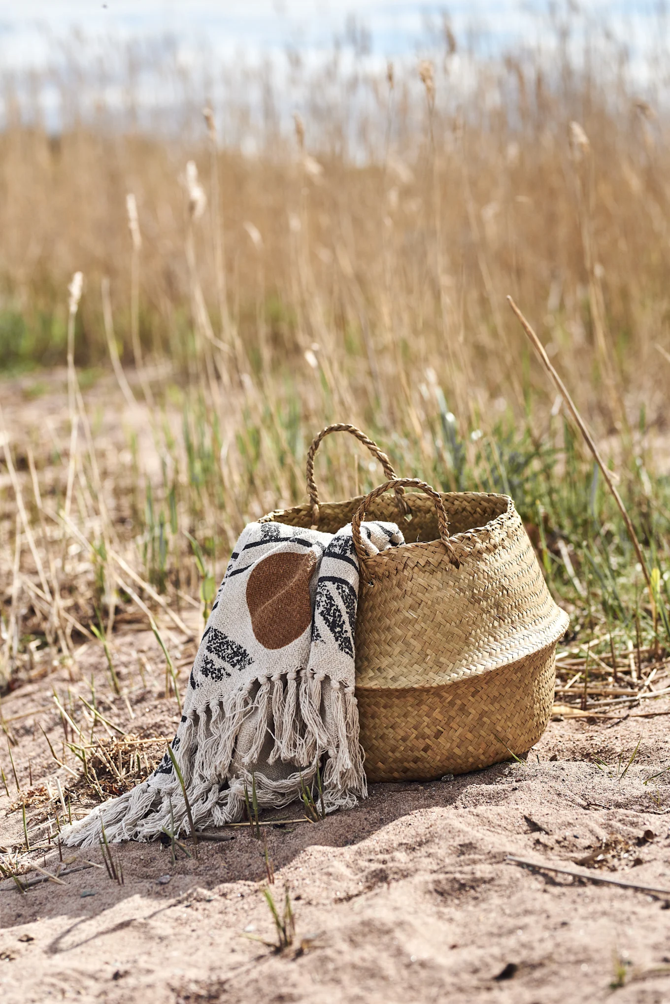 Un panier en paille tressée avec une couverture blanche à motifs sur le sable, entouré de roseaux secs.