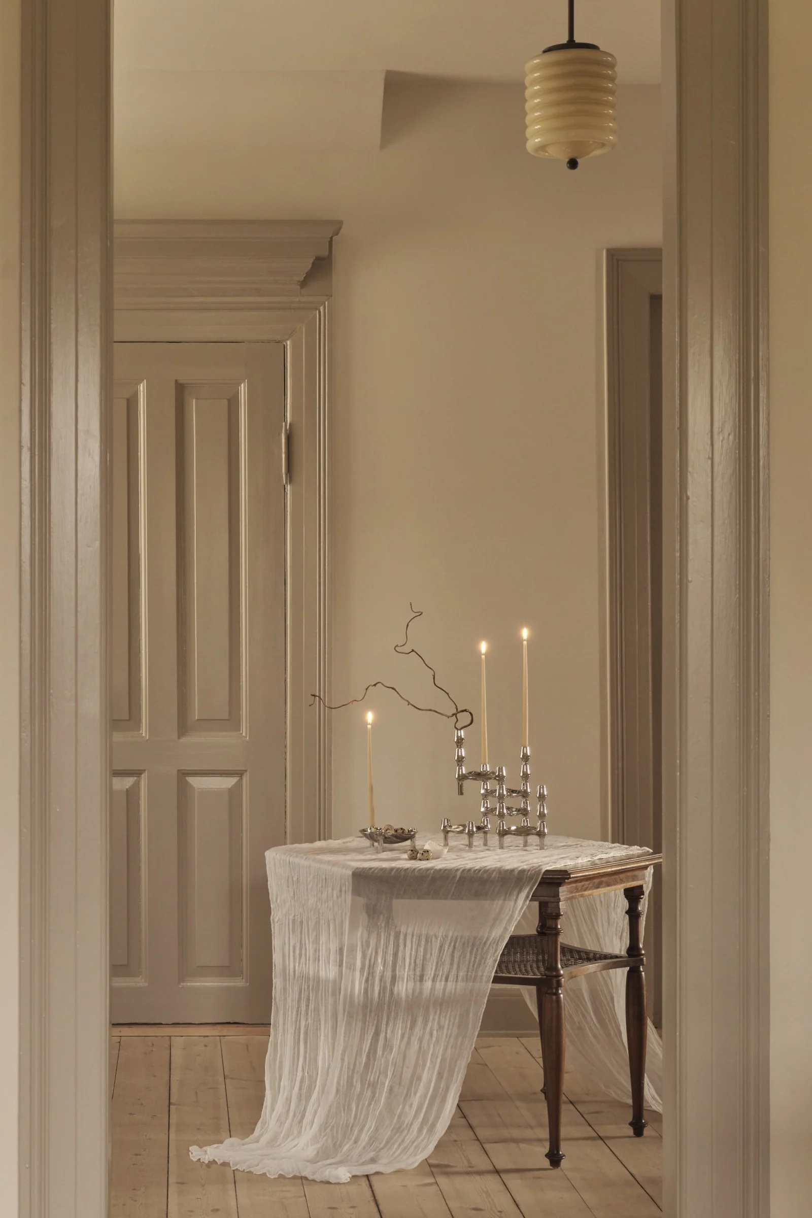 Dans une pièce aux tons sobres, une table en bois recouverte d'une nappe blanche transparente accueille trois bougies allumées dans des chandeliers en argent, un bol d'œufs mouchetés et une branche.