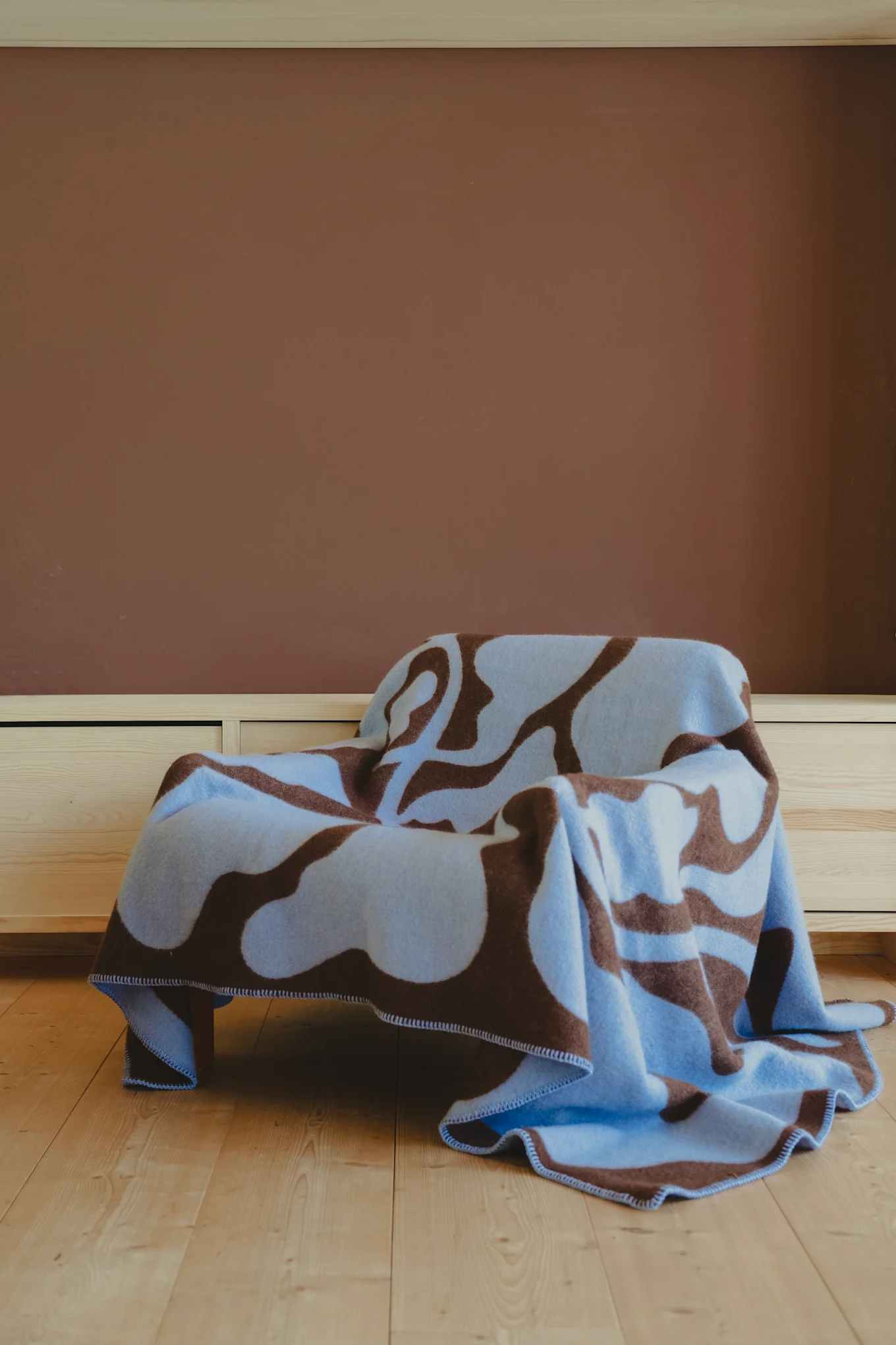 La couverture Leaves à motifs bruns et bleus de Fine little day est posée sur un fauteuil et fait partie des tendances 2025 de la décoration d'intérieur grâce à sa palette de couleurs.