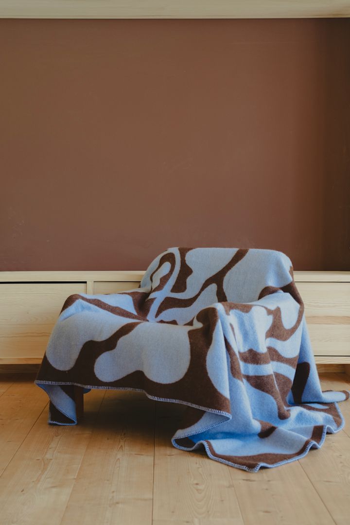 La couverture Leaves à motifs bruns et bleus de Fine little day est posée sur un fauteuil et fait partie des tendances 2025 de la décoration d'intérieur grâce à sa palette de couleurs.