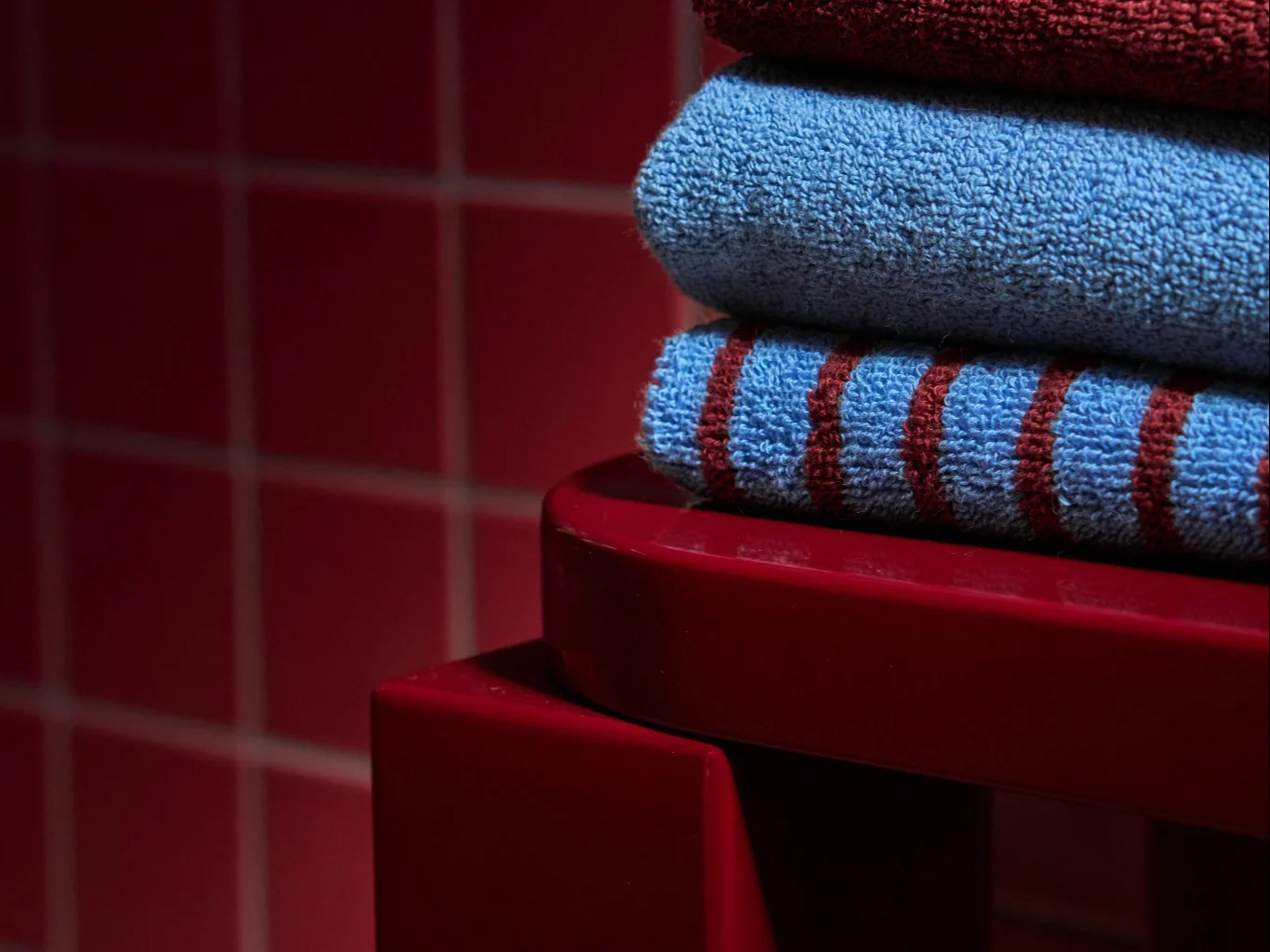 Pile de trois serviettes pliées : rouge foncé, bleu clair et bleu à rayures rouges, sur une surface rouge.