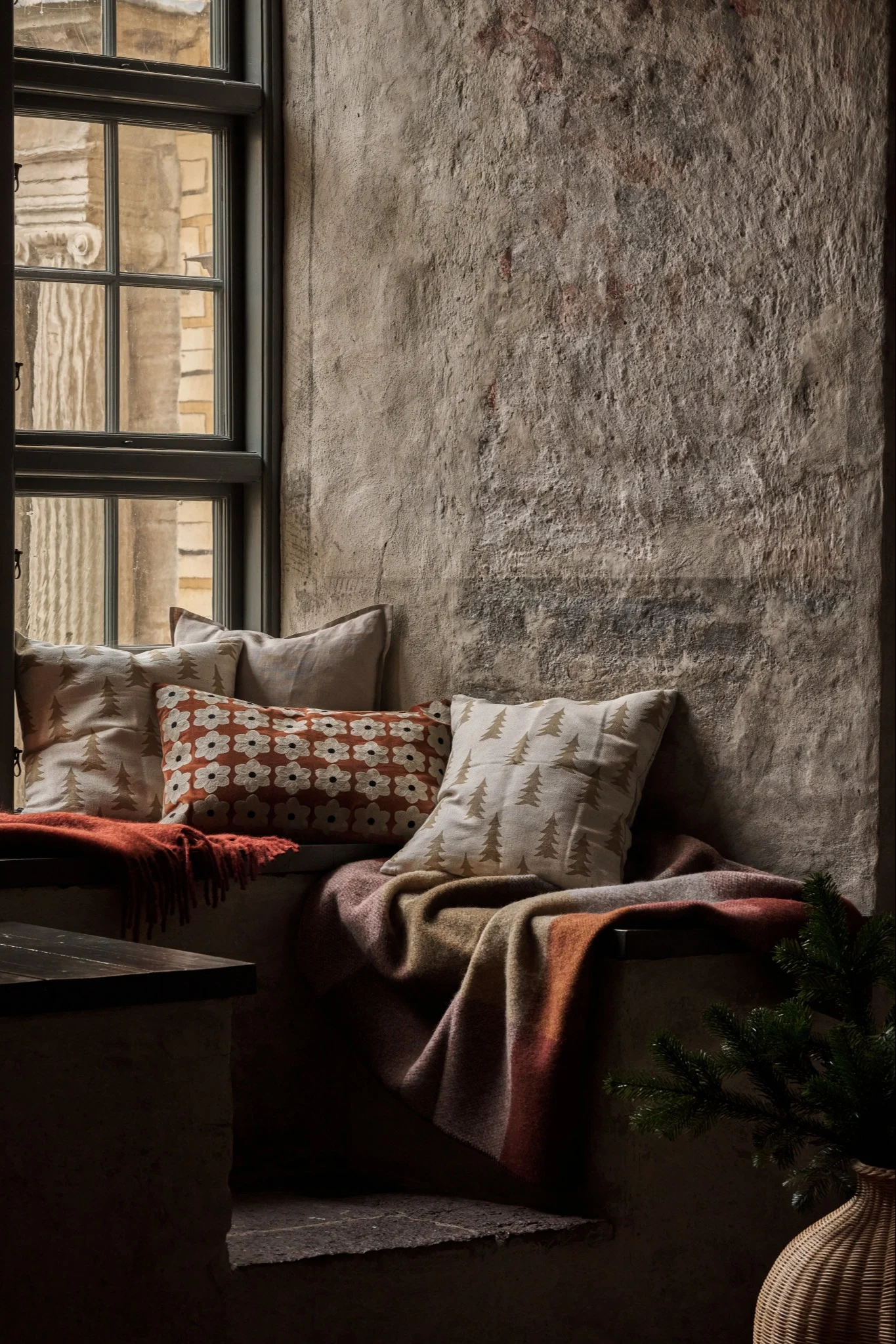 Banquette de fenêtre confortable avec coussins à motifs, couvertures chaudes et branche de pin près d'un mur rustique.