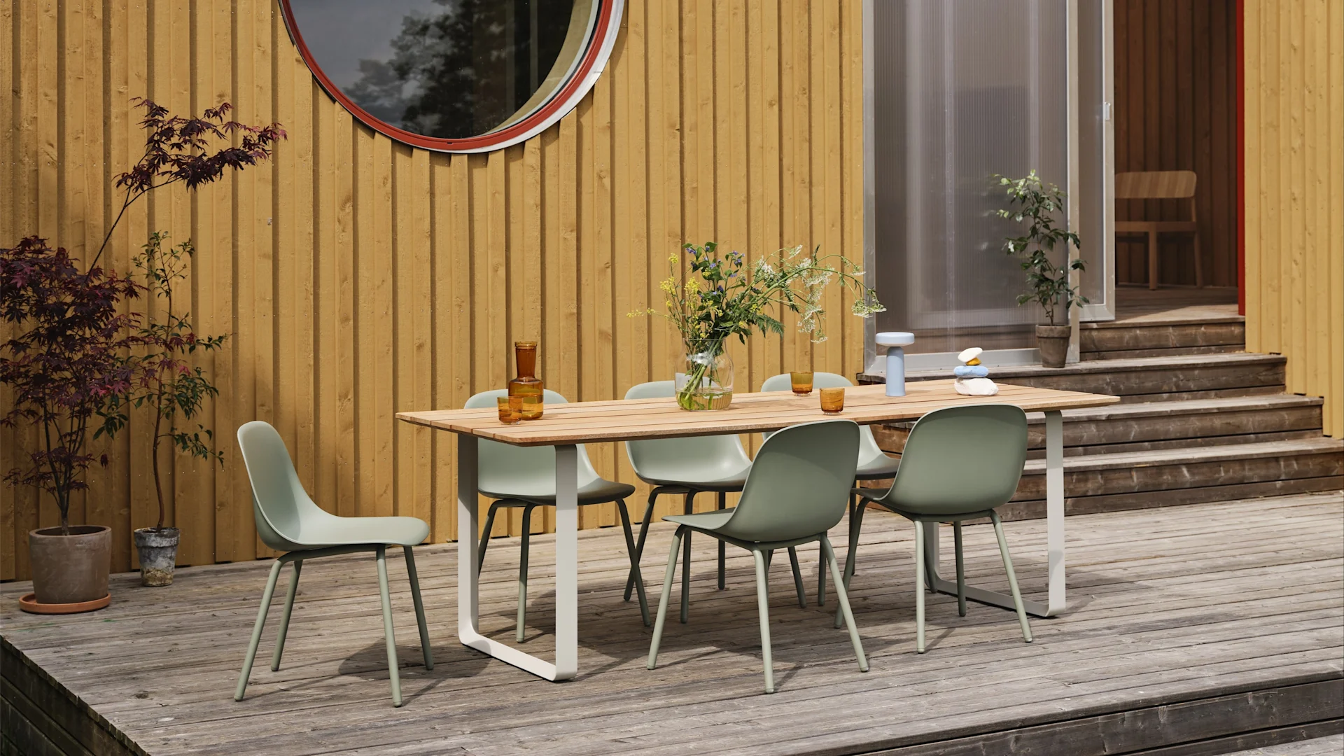Tendances meubles de jardin 2025 : les chaises d'extérieur vert sauge Fiber de Muuto sont disposées sur une grande terrasse et autour d'une table, avec en arrière-plan une maison en bois jaune.