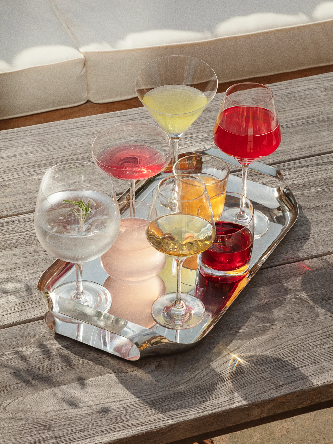 Un plateau en argent avec diverses boissons colorées sur une table en bois, avec un canapé blanc en arrière-plan, baigné de soleil.