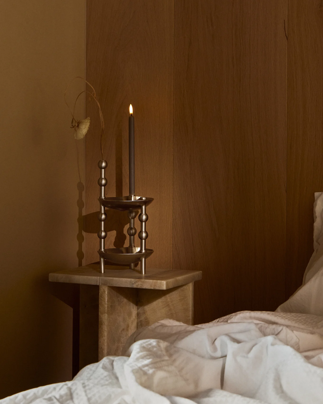 Une bougie sombre allumée dans un bougeoir métallique orné sur une table de chevet en bois, à côté d'un lit avec une literie blanche dans une pièce sombre.