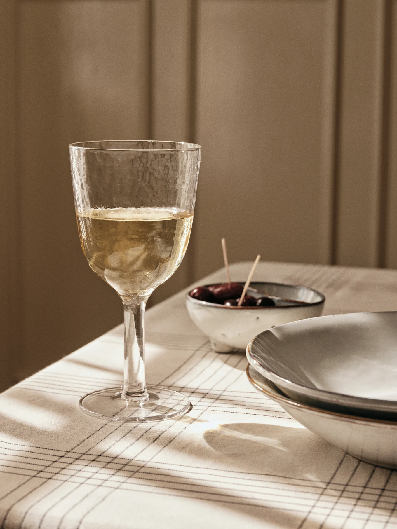 Un verre de vin blanc texturé sur une nappe rayée, avec des olives et des assiettes en arrière-plan, baigné par la lumière de la fenêtre.