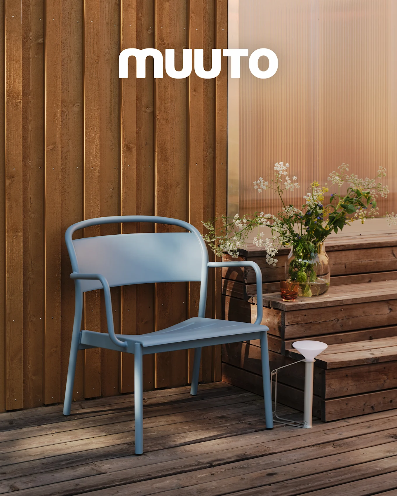 Une chaise d'extérieur bleu clair sur une terrasse en bois près de marches avec un vase de fleurs sauvages et une lampe portable blanche. Logo MUUTO.