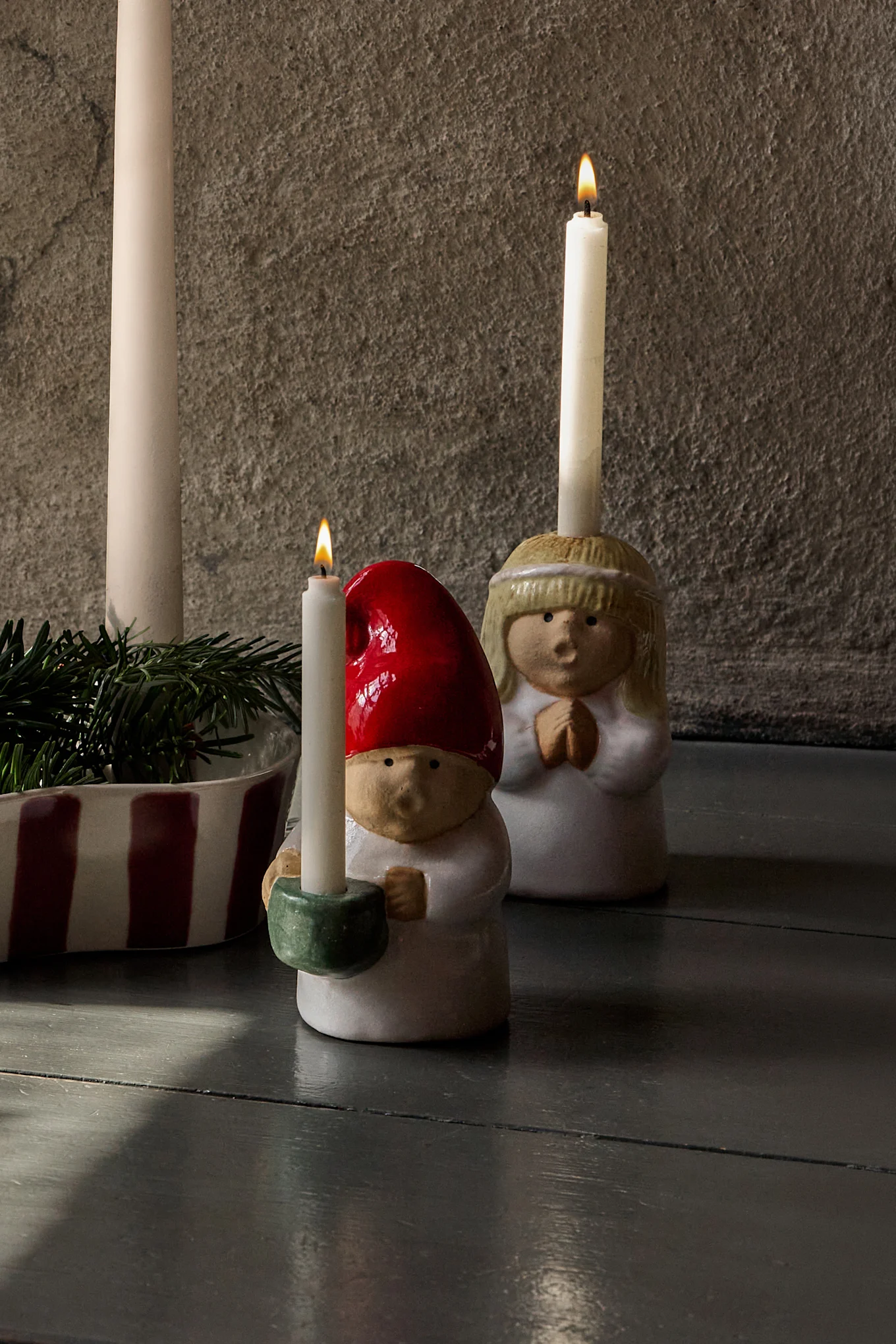 Arrangement de l'Avent : quatre bougies éteintes dans un support rayé avec du pin, flanquées de figurines en céramique (ange et gnome) avec des bougies allumées.