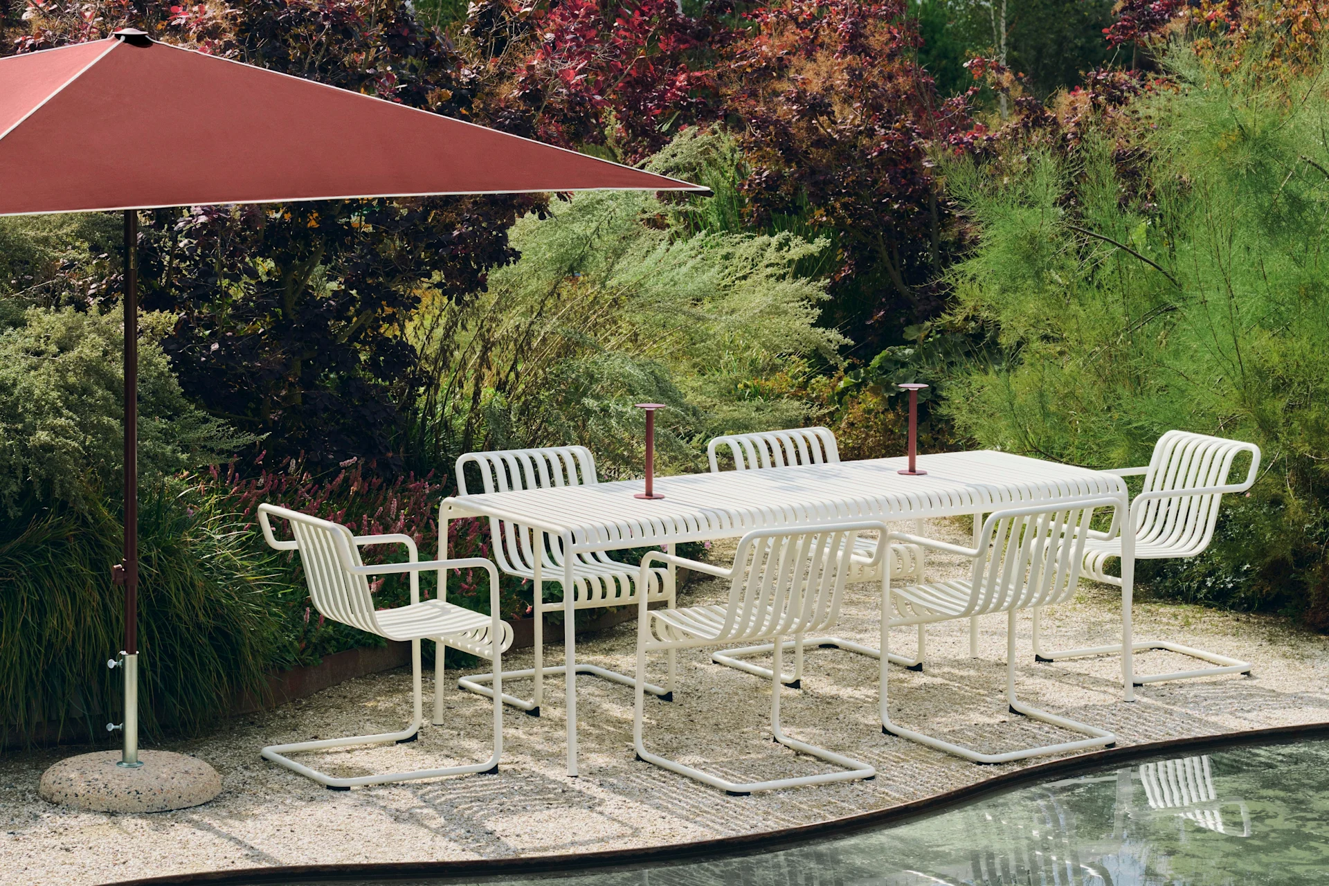 Ensemble de repas extérieur avec table à lattes blanche et six chaises sous un grand parasol rouge, sur patio de gravier près d'un étang.