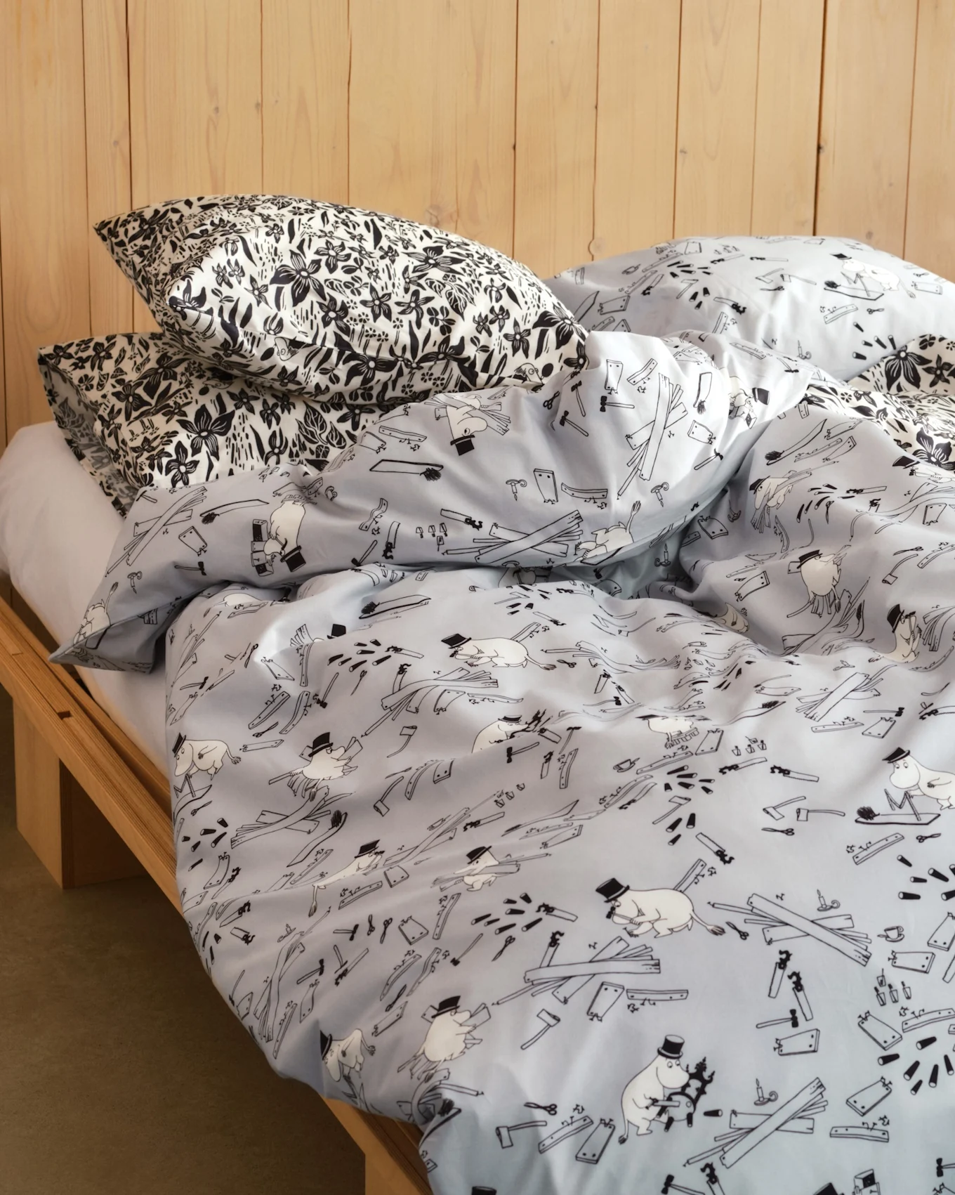 Lit avec oreillers à motif floral noir et blanc et housse de couette bleu clair avec figures de dessins animés, outils et planches de bois. Cadre de lit en bois.