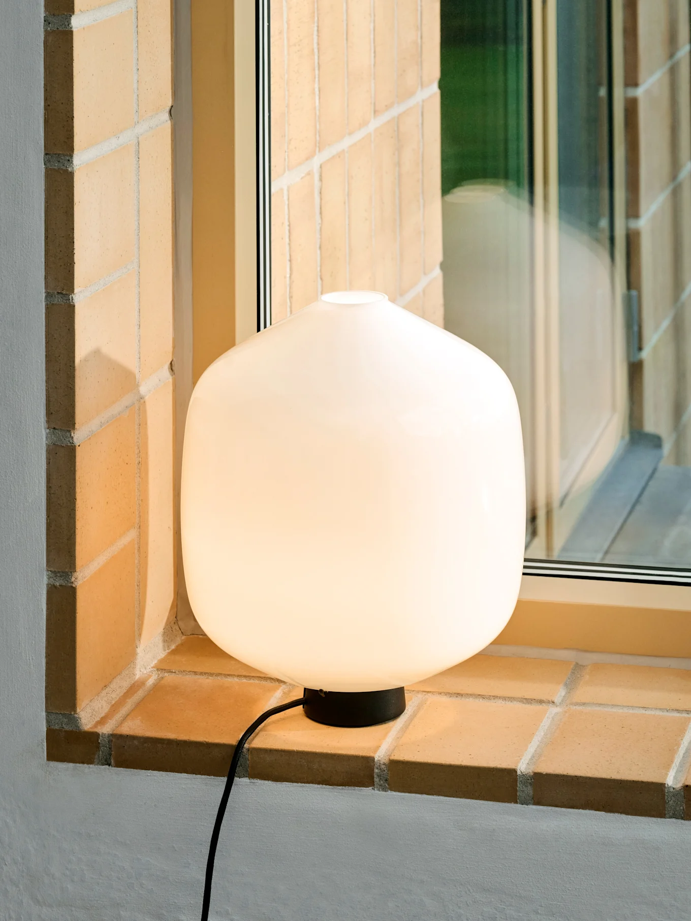 Une lampe de table blanche, lumineuse et arrondie avec une base et un cordon noirs sur un rebord de fenêtre en brique.