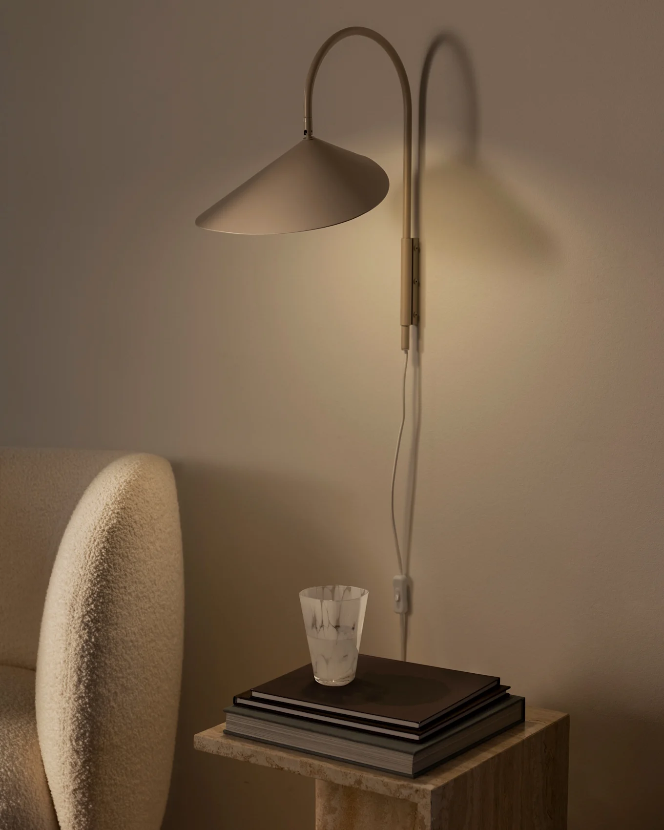 Lampe murale chaleureusement éclairée avec abat-jour conique au-dessus de livres et verre texturé sur table en pierre, à côté d'un fauteuil beige.
