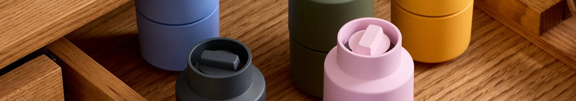 Cinq récipients colorés : gris foncé, bleu clair, olive, rose clair avec un capuchon unique et jaune moutarde, sur bois.