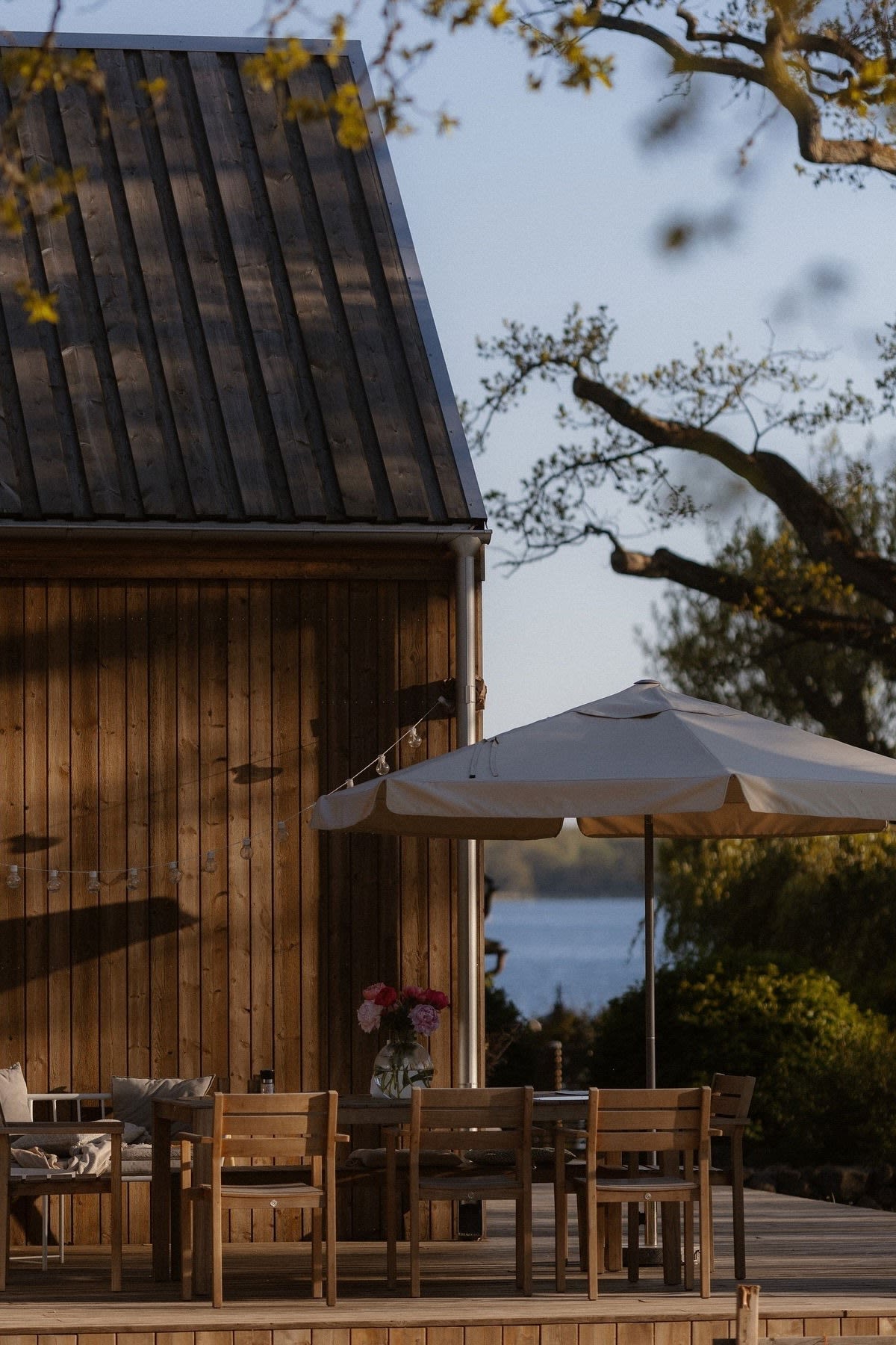 Un patio confortable en face d'une maison en bois avec un coin repas avec des chaises Sköndal de 1898 et un parasol.
