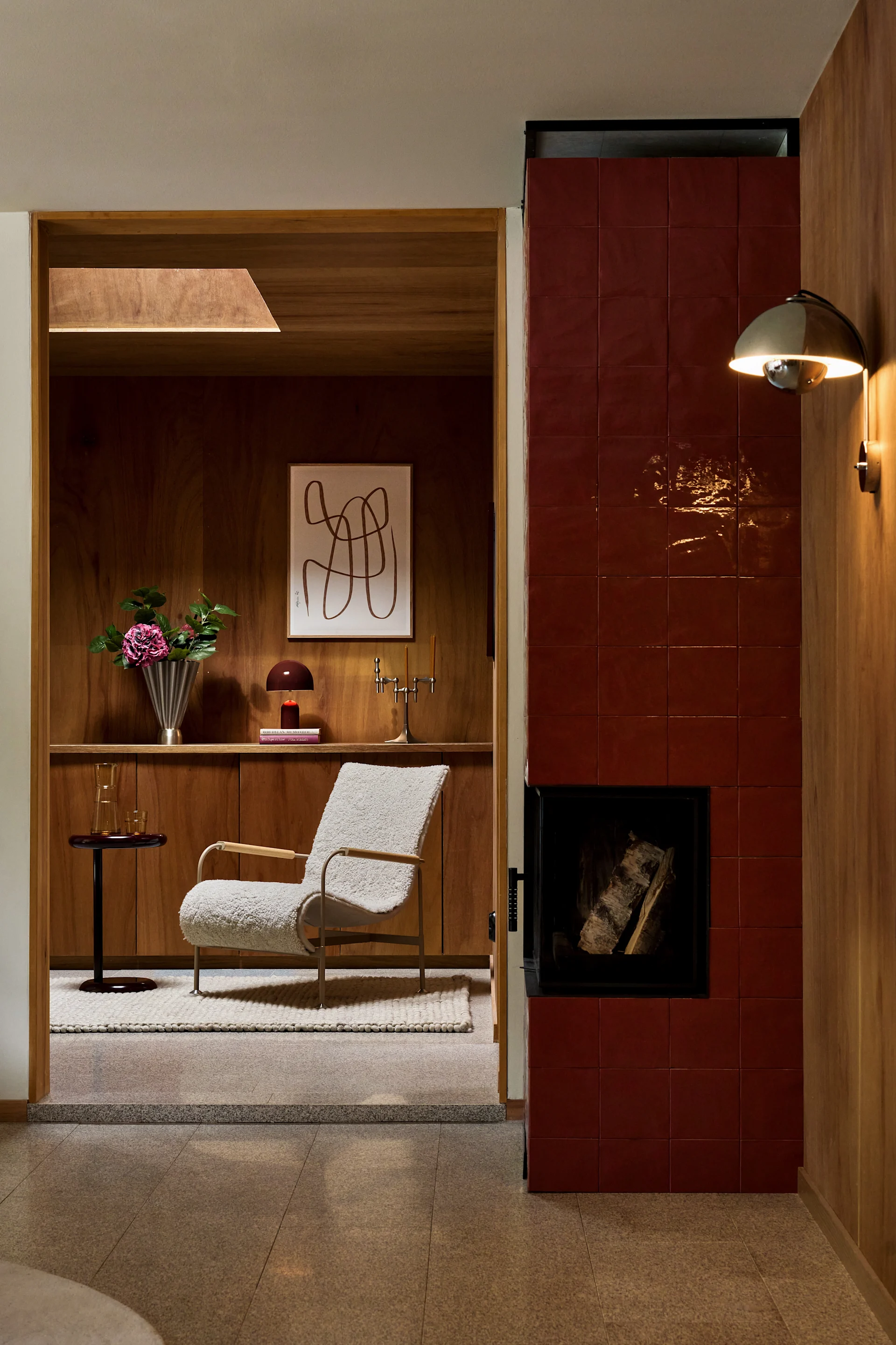 Un espace de vie confortable avec une cheminée carrelée rouge, un fauteuil bouclé crème et des panneaux muraux en bois avec étagères intégrées.
