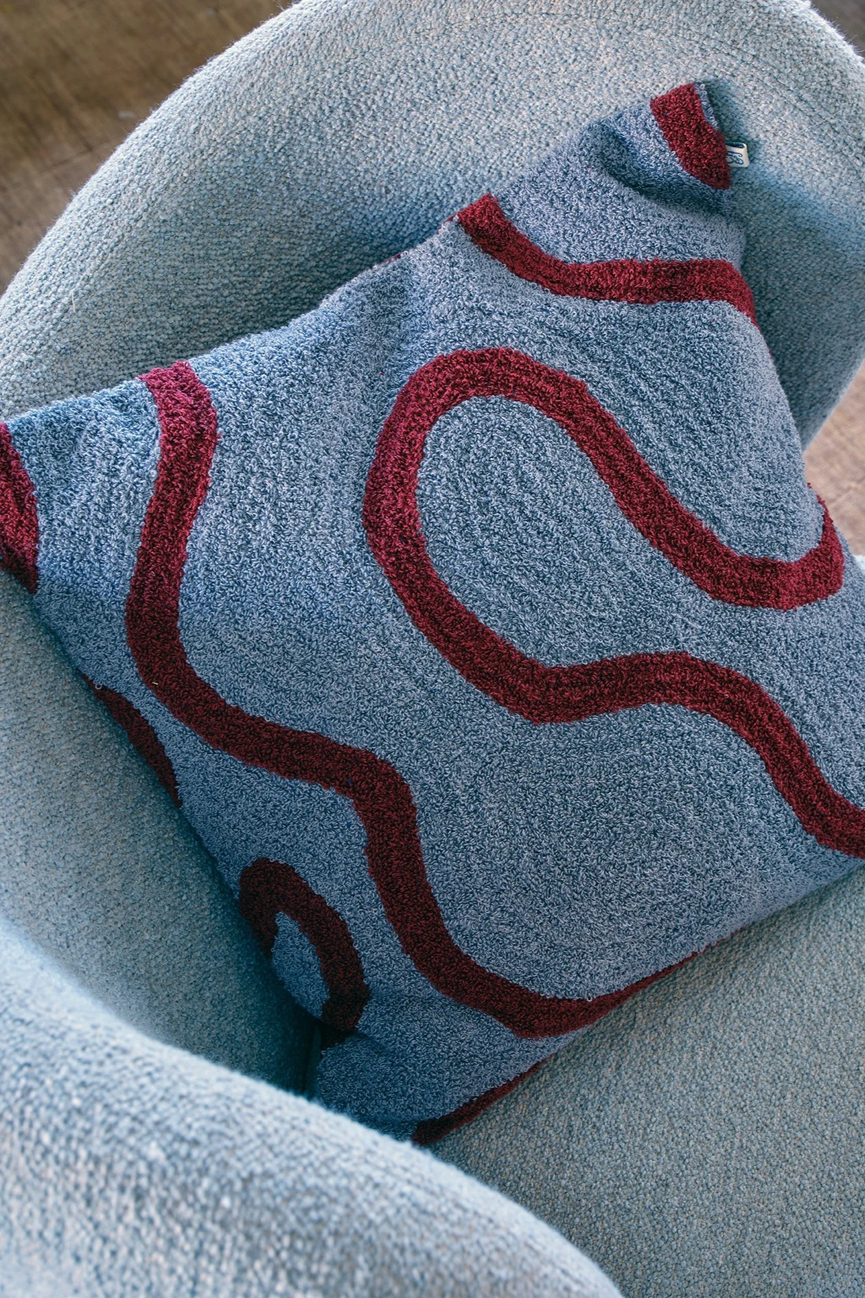 Un coussin bleu clair texturé, orné d'un motif ondulé rouge foncé, posé sur un fauteuil bleu clair.