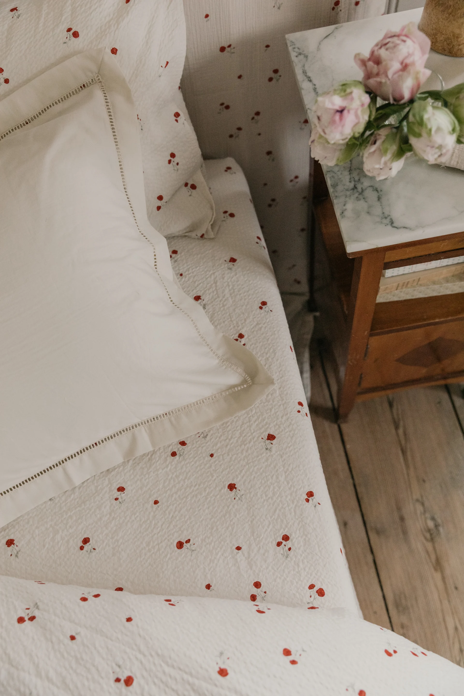 Literie blanche à motif floral rouge, un oreiller blanc et des fleurs roses sur une table de chevet en marbre.