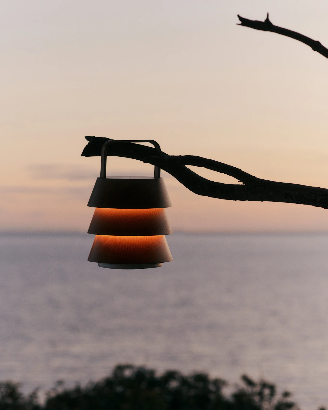 Lanterne à abat-jour étagés suspendue à une branche devant un ciel de coucher de soleil sur l'eau.