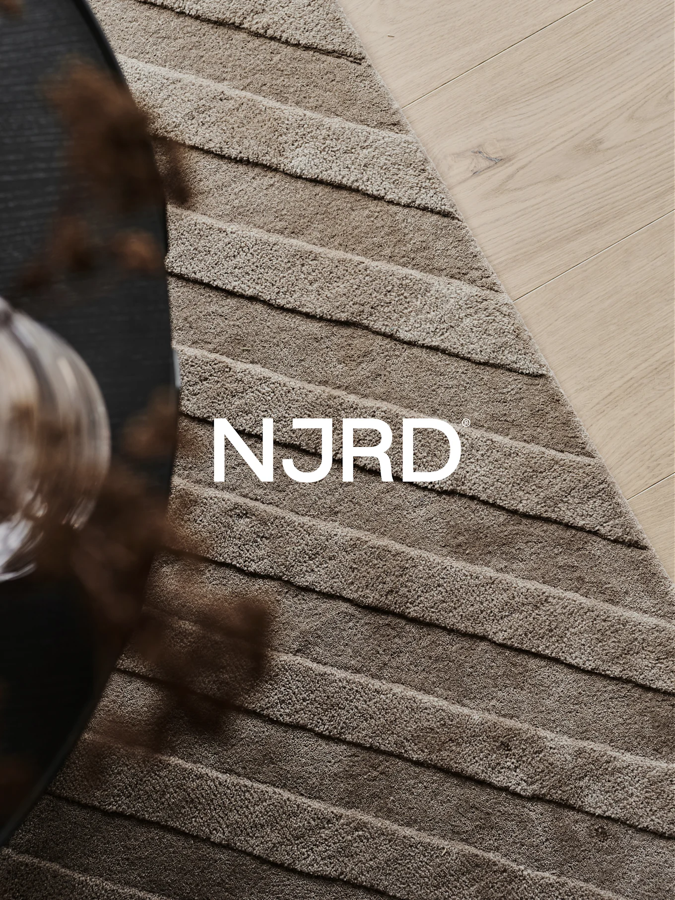 Gros plan d'un tapis shaggy marron clair à rayures horizontales, partiellement posé sur un parquet en bois clair, avec le logo blanc 'NJRD'.