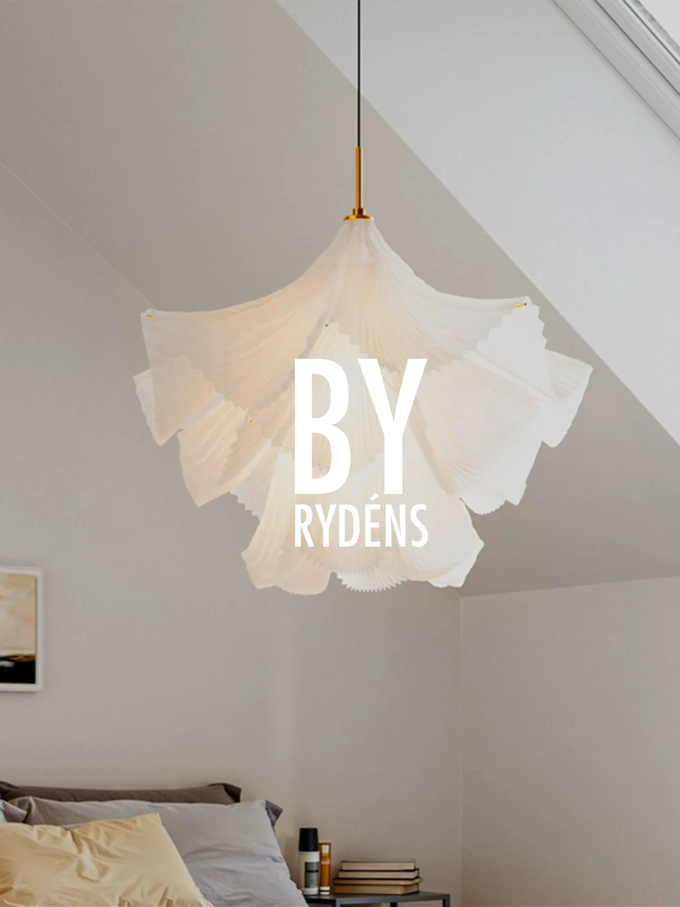 Une suspension sculpturale plissée blanche est suspendue au-dessus d'un lit avec des oreillers et une table de chevet. 'BY RYDÉNS' est superposé sur la lampe.