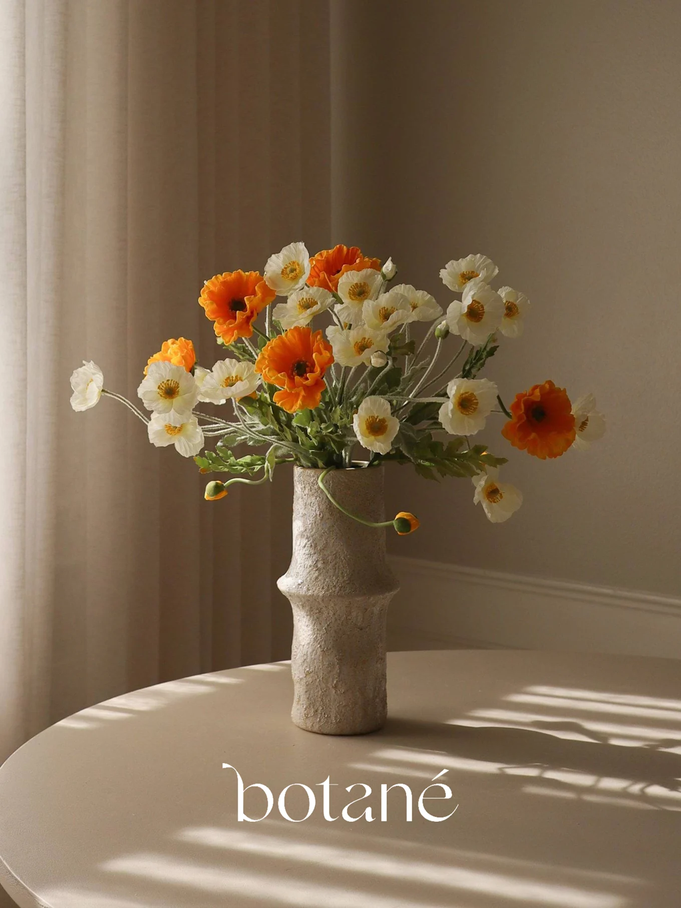 Un vase texturé contient un bouquet de coquelicots artificiels oranges et blancs sur une table aux ombres rayées.