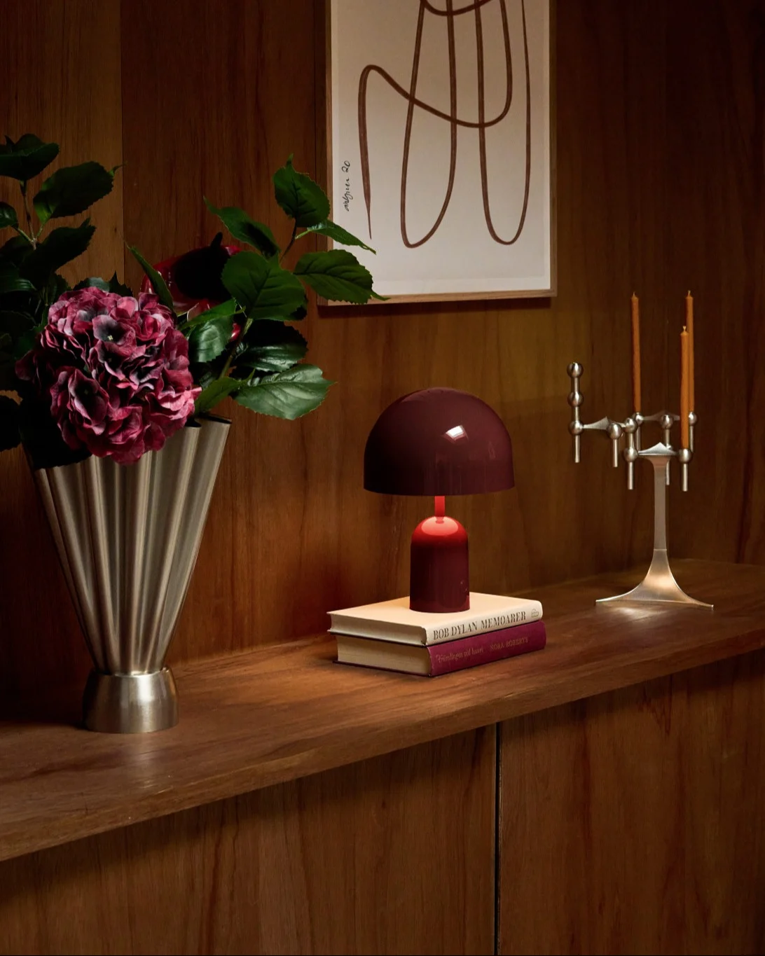 Étagère en bois avec vase cannelé argenté, hortensias rouges, lampe rouge sur livres et candélabre argenté.